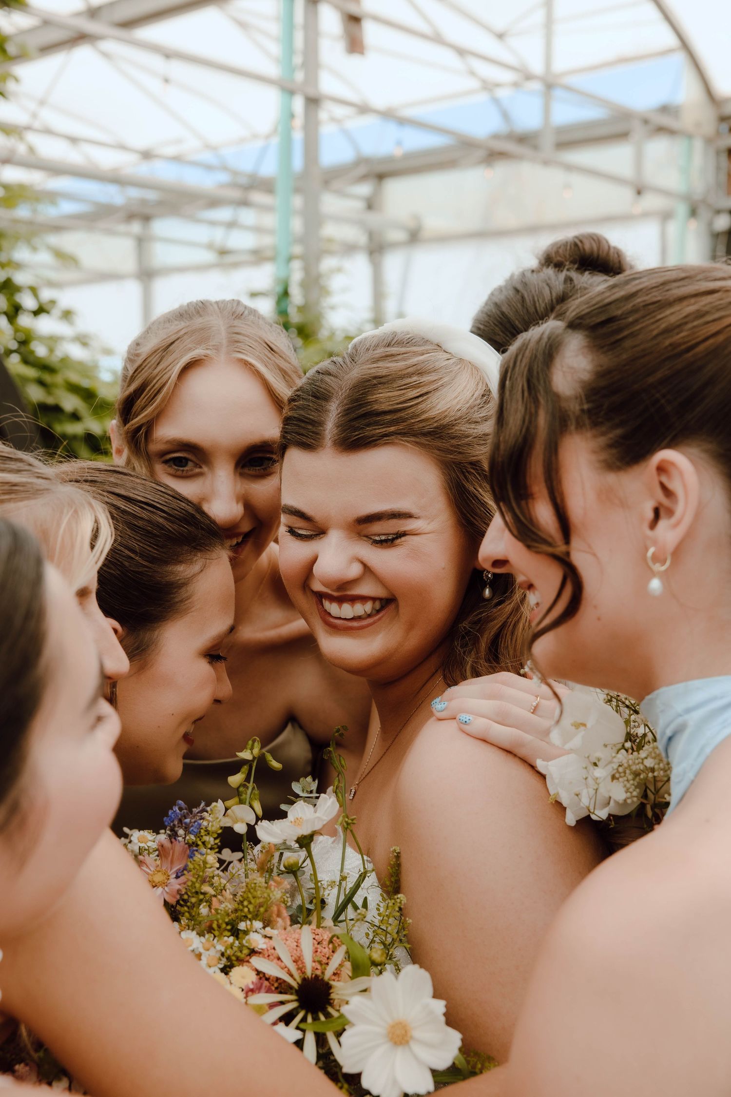 Bridesmaids huddling together in a greenhouse, laughing and holding wildflower bouquets at a wedding celebration.