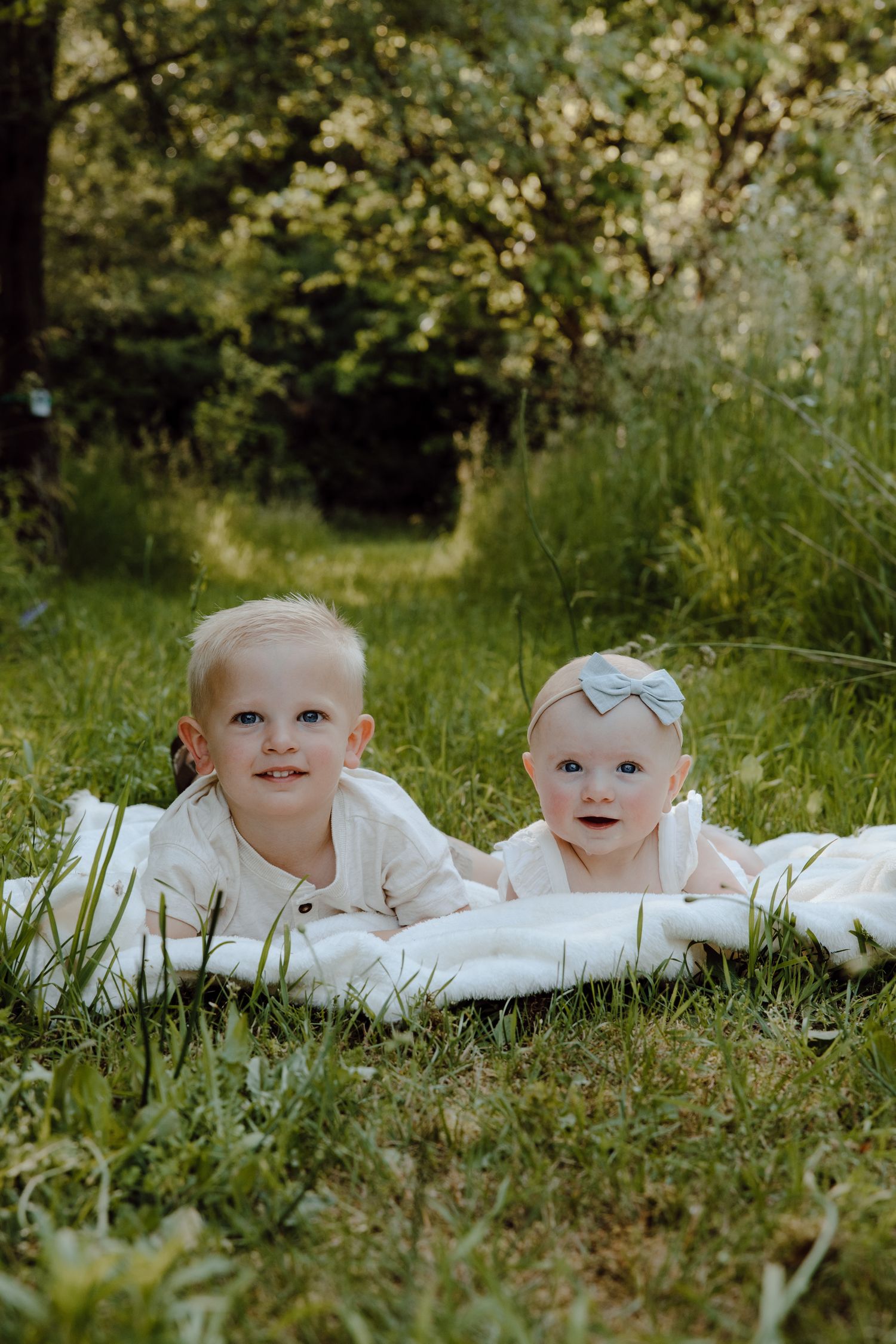 Two young children in white outfits lying on a blanket in green grass during a sunny outdoor photoshoot.