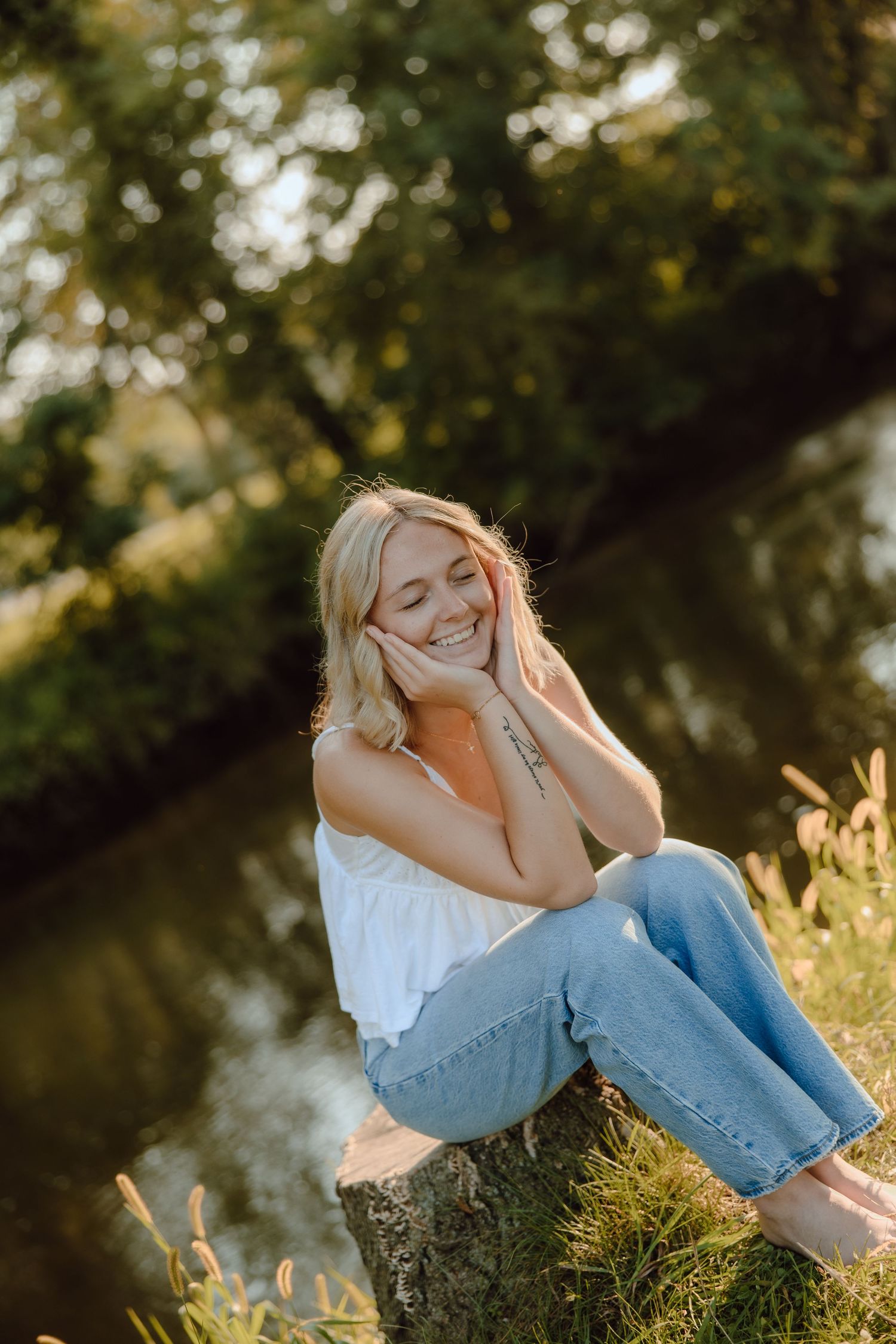 Smiling blonde woman sitting on a tree stump by a pond in golden sunlight, wearing white top and blue jeans.