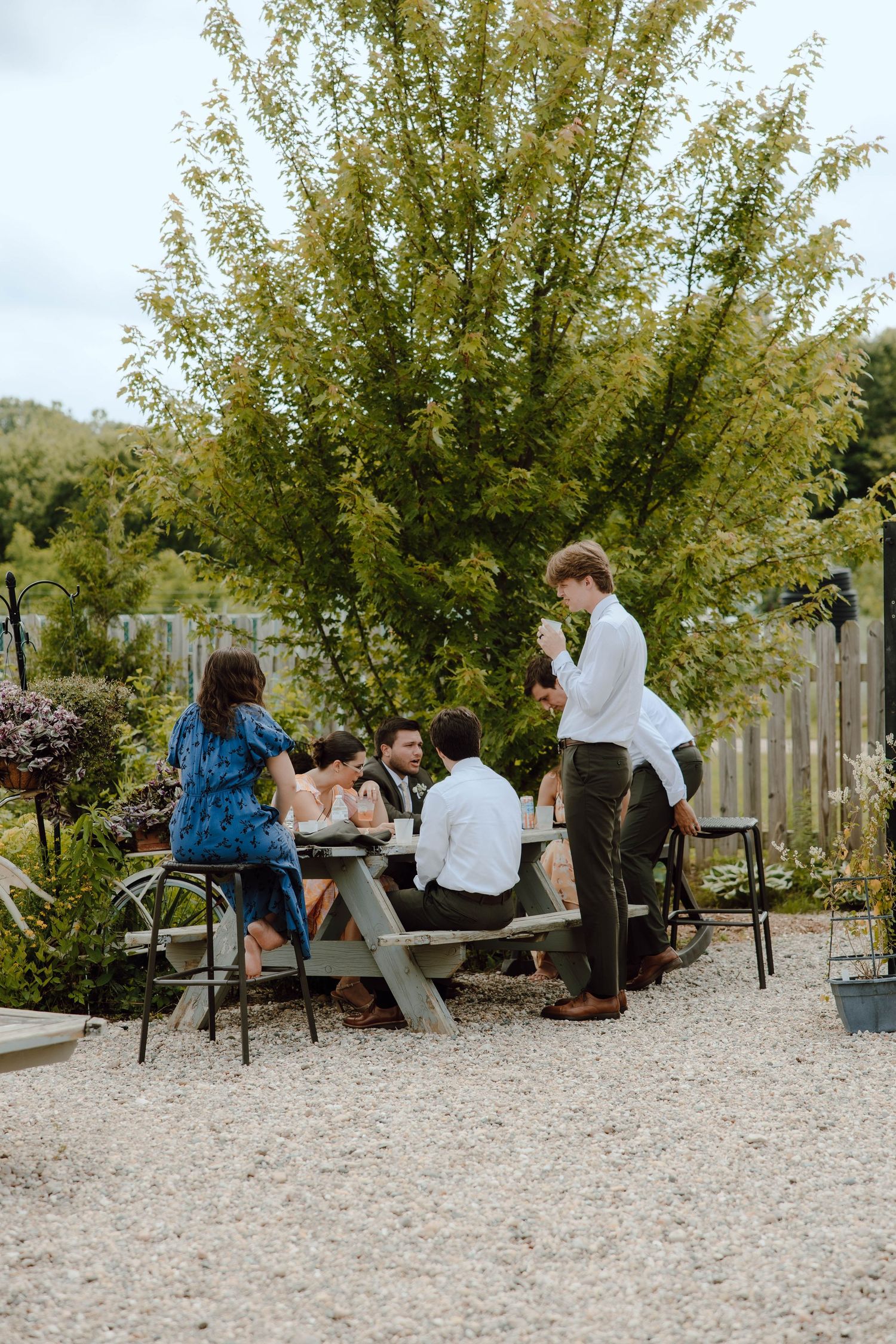 Wedding guests gather around picnic table in garden setting during outdoor wedding reception.