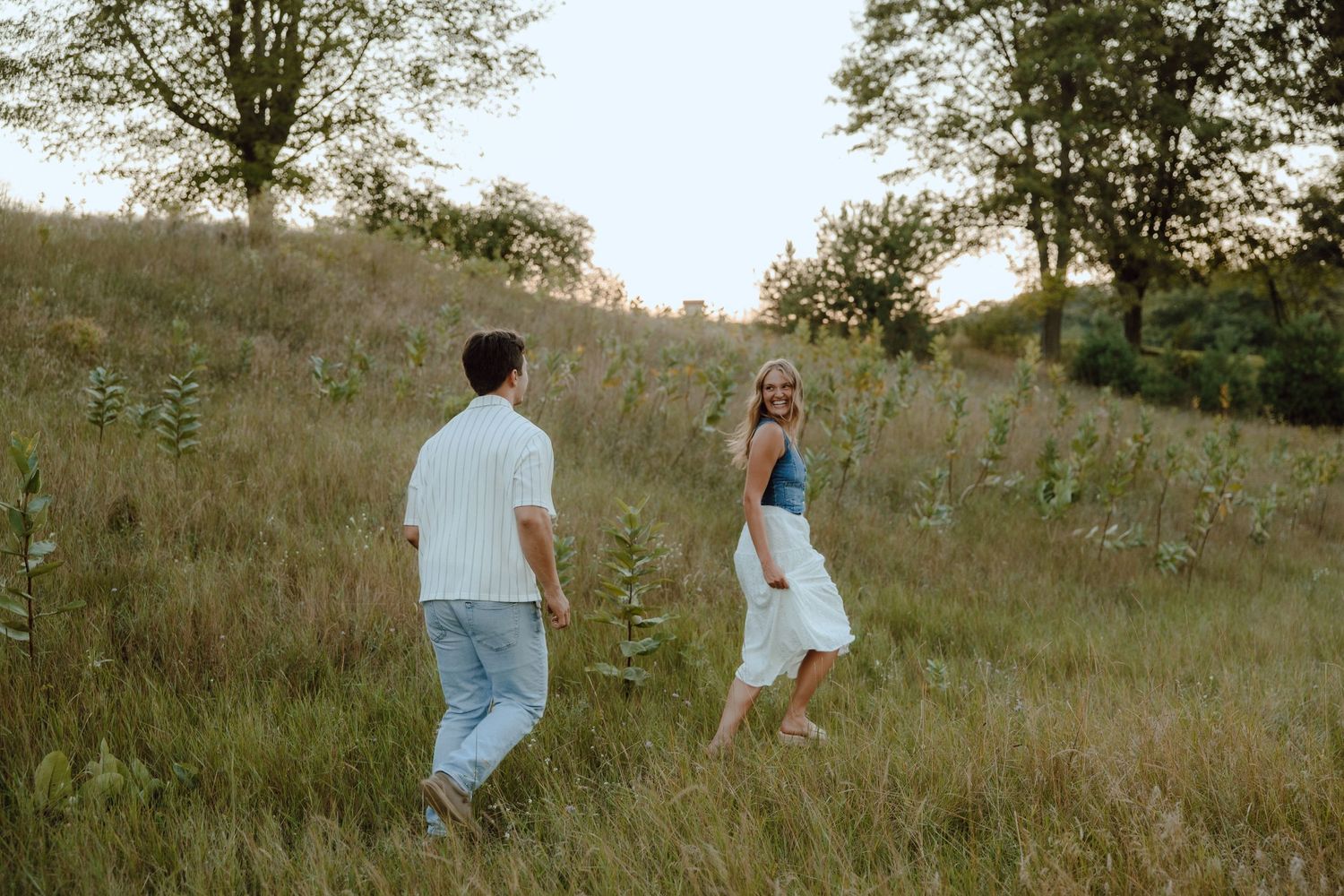 Couple walking through a lush green meadow at golden hour, woman in white skirt smiling back.