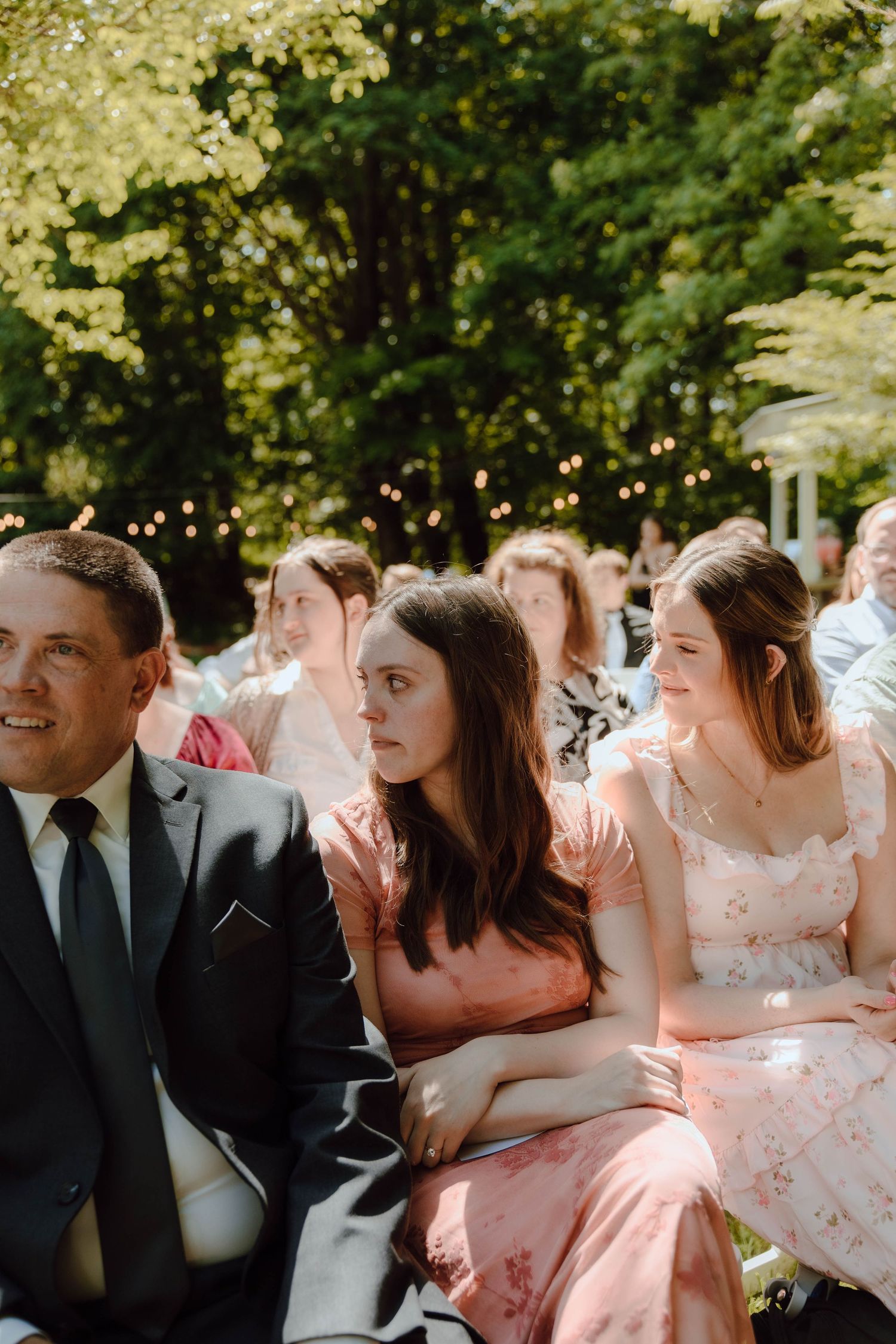 Wedding guests seated outdoors in sunlight watch the ceremony, surrounded by trees and string lights.