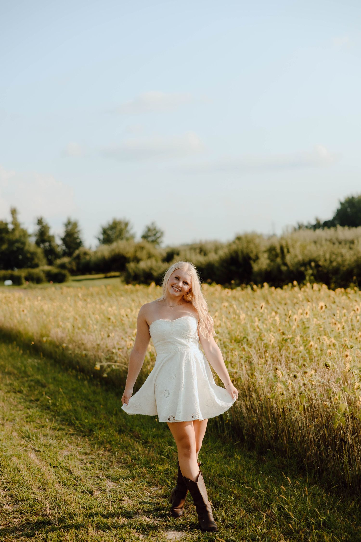 Blonde woman in white strapless dress and cowboy boots walking along a sunny field path.