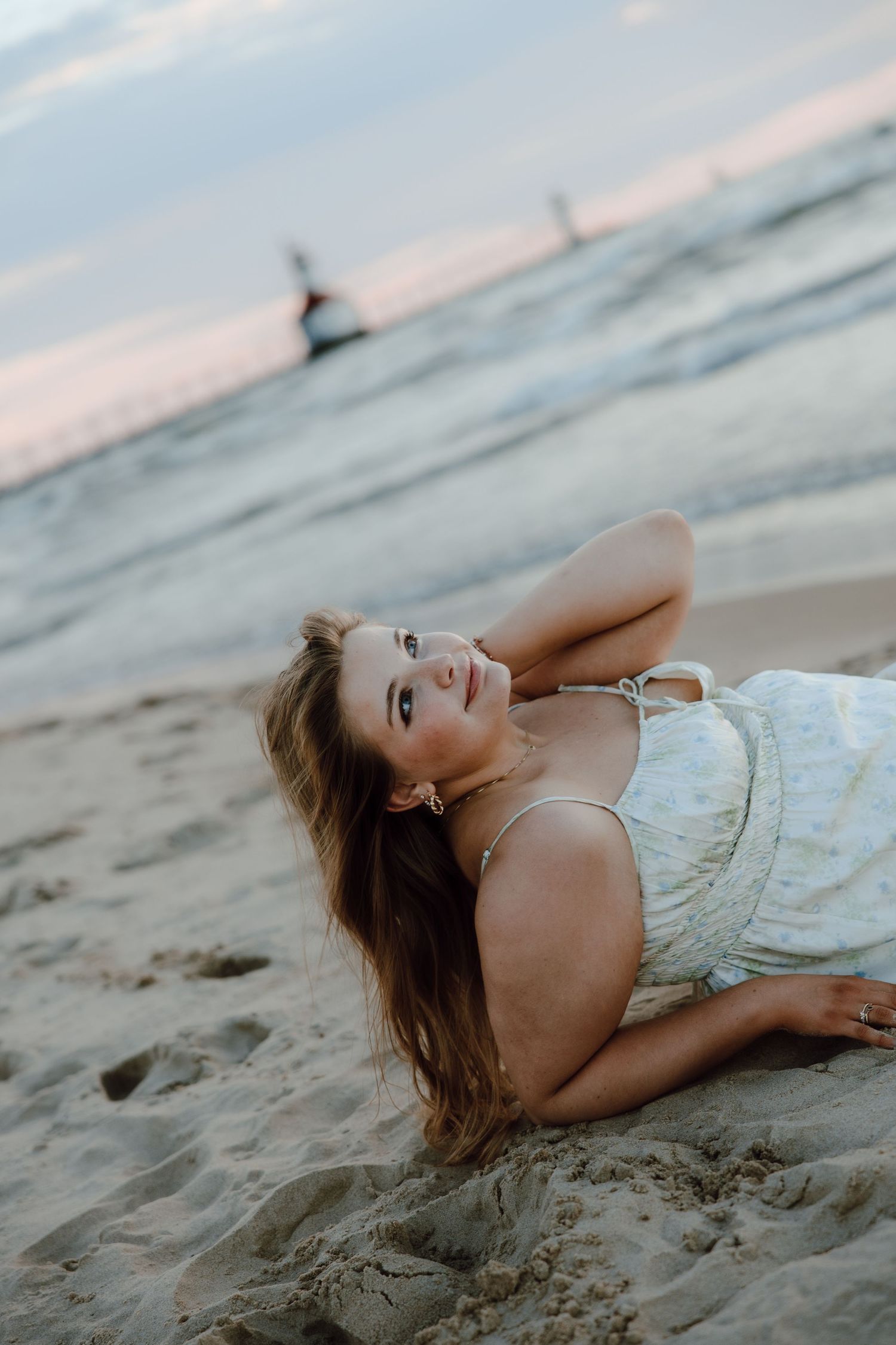 Young woman in white dress lying on sandy beach at sunset, smiling with long hair flowing, ocean waves in background.