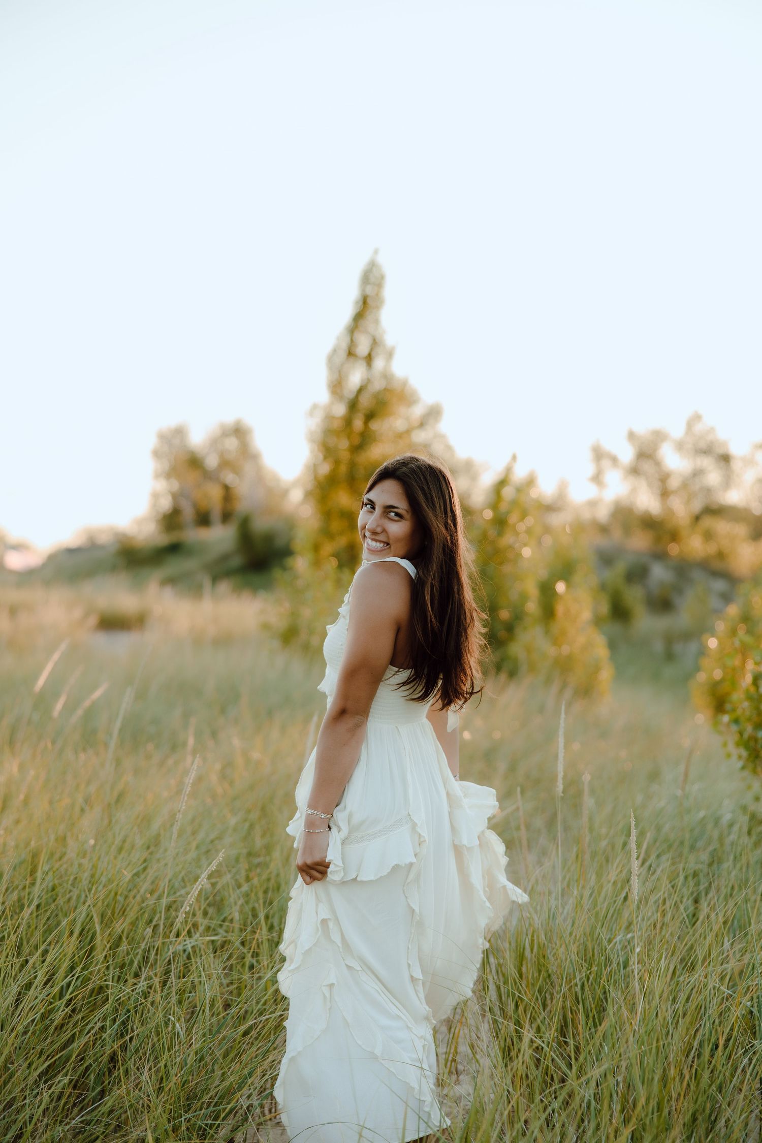 A brunette woman in a flowing white dress stands in a golden field, smiling over her shoulder at sunset.