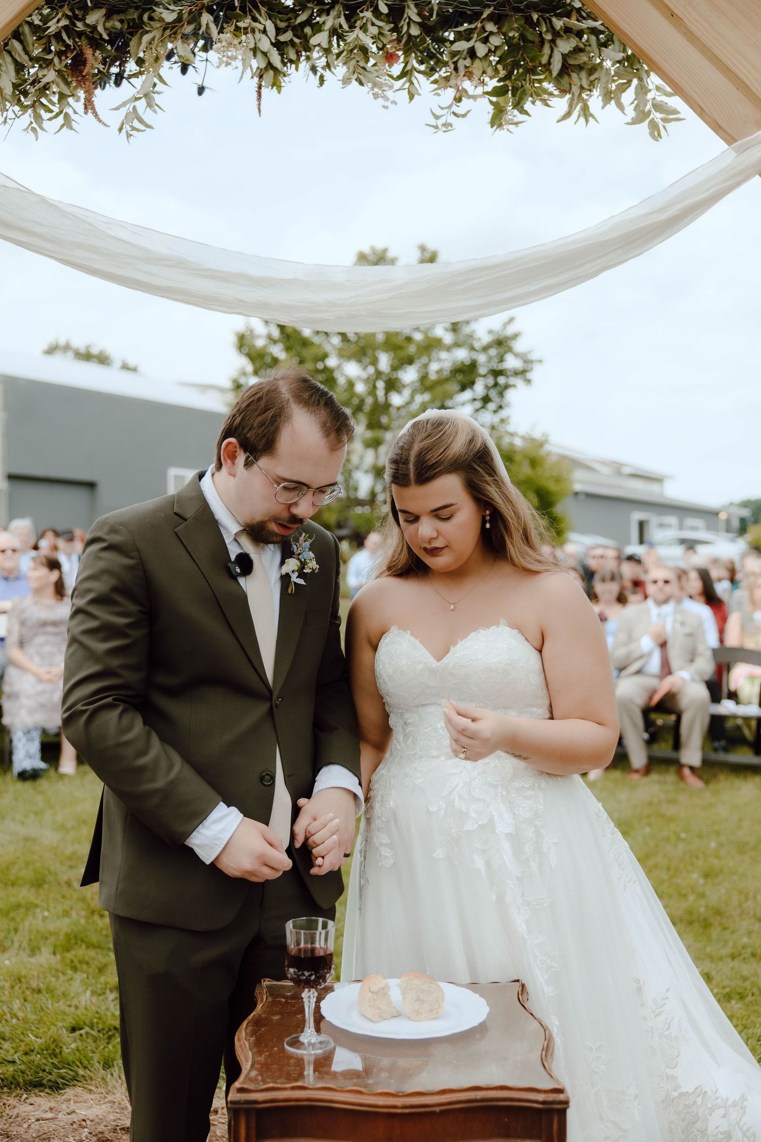 Bride and groom perform a unity ceremony outdoors under a floral arch, with wedding guests seated in the background.
