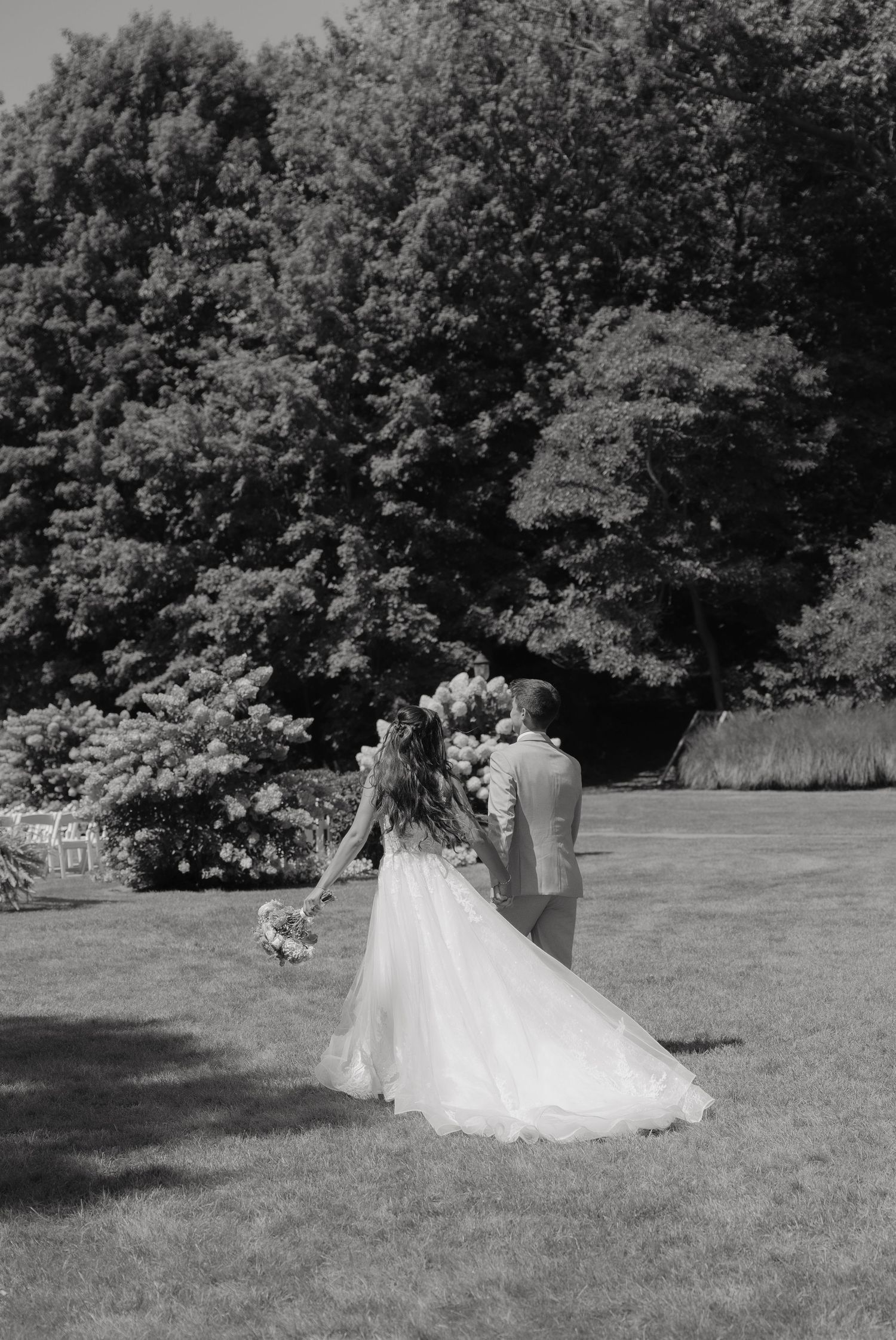 Black and white photo of a bride and groom walking hand in hand across a lush green lawn toward flowering trees.