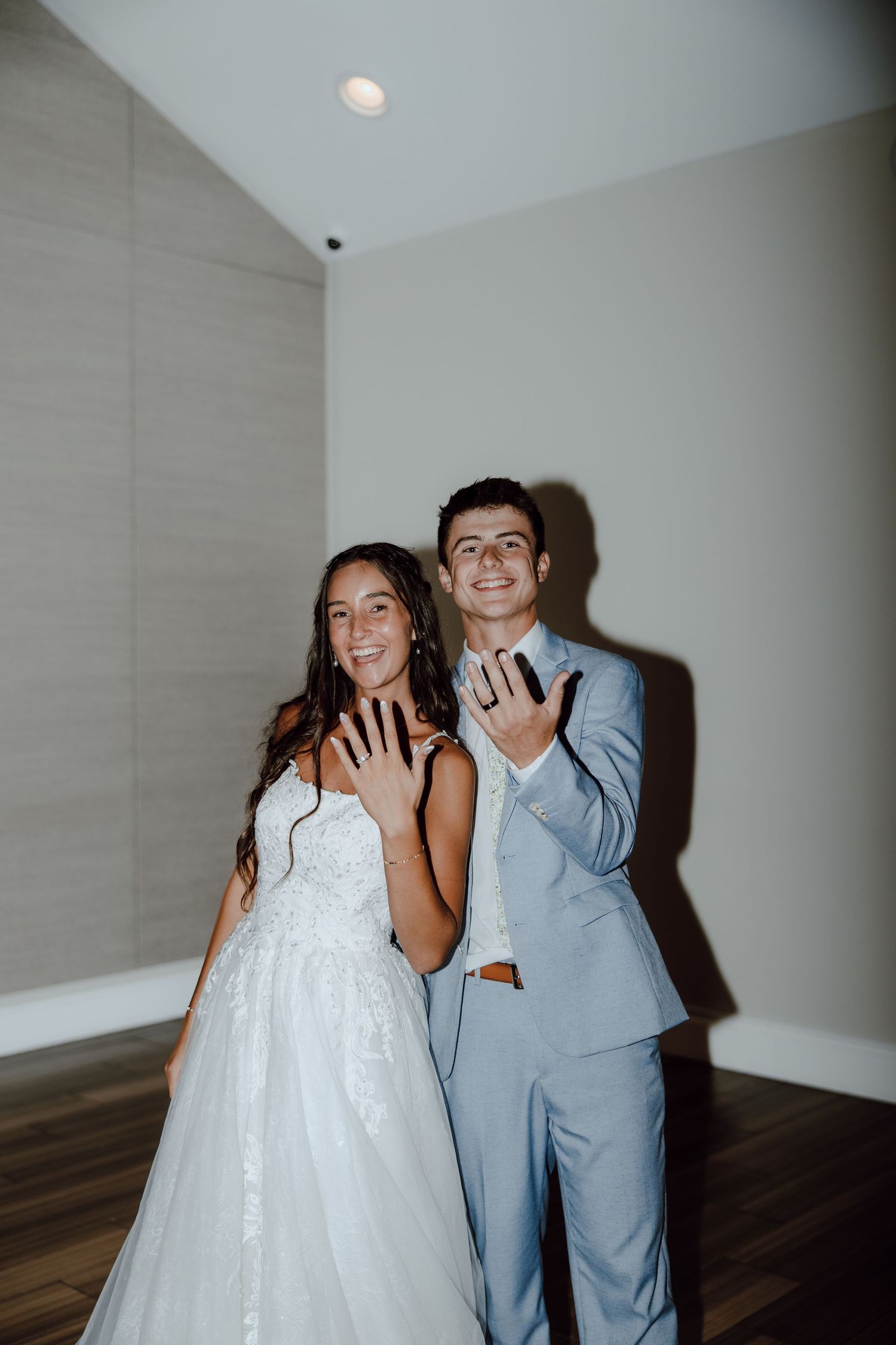 Smiling bride in white lace gown and groom in light blue suit showing off their wedding rings indoors.