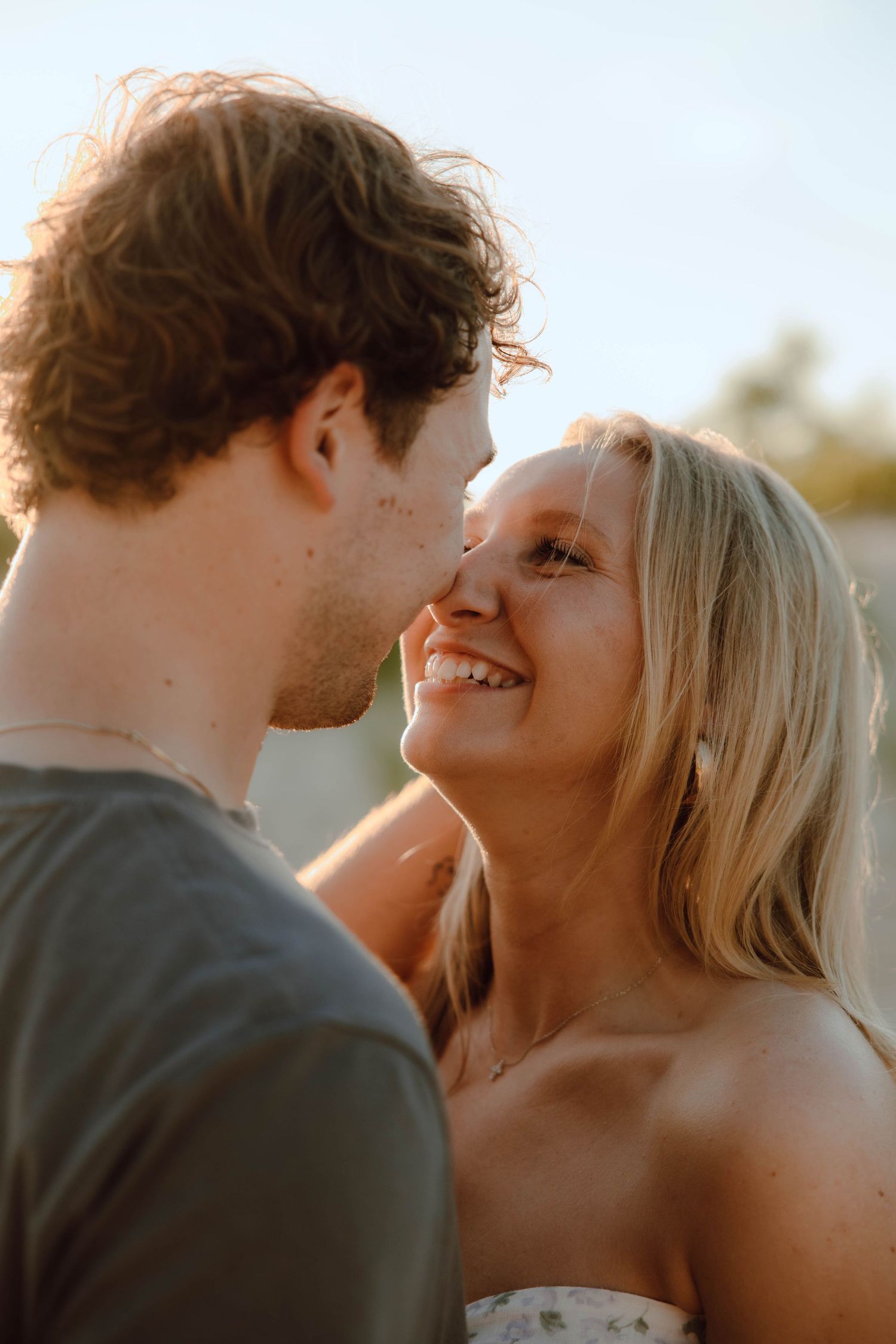 A smiling blonde woman and curly-haired man share a romantic moment outdoors in golden hour light.