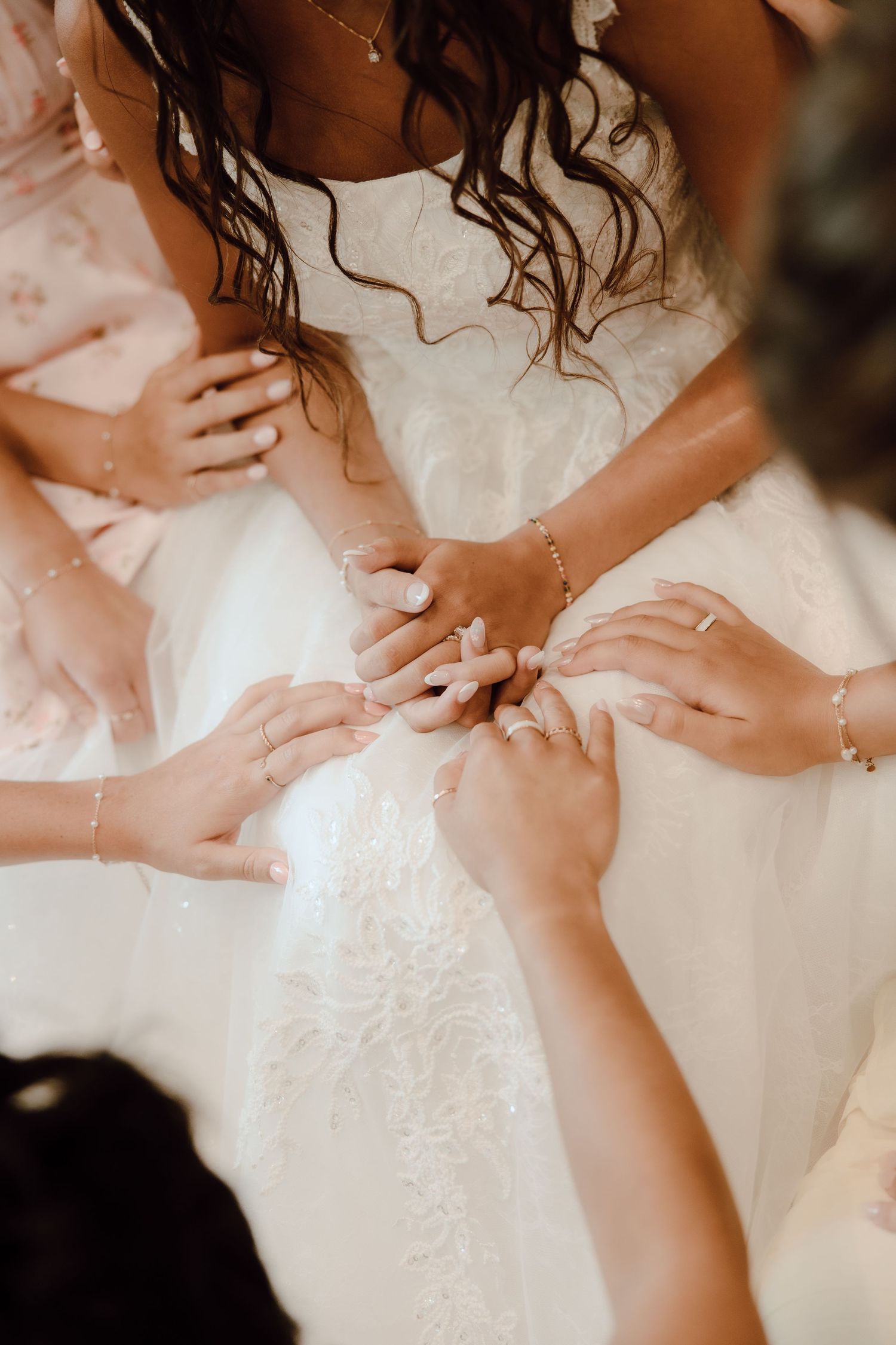 Bride in white lace gown with bridesmaids holding hands together in a touching wedding preparation moment.