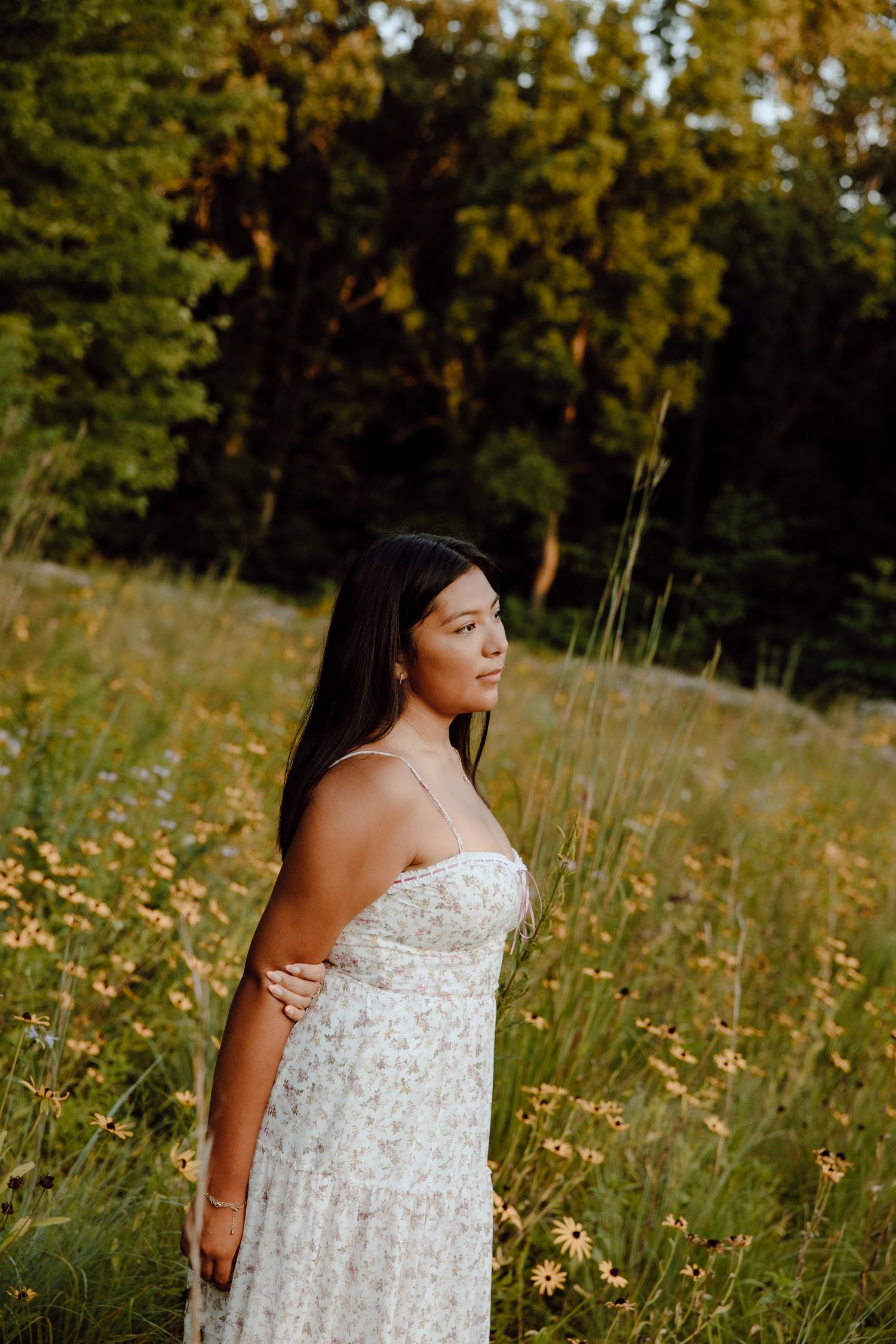 Elegant poses in a white sundress amid wild grasses and flowers at sunset.