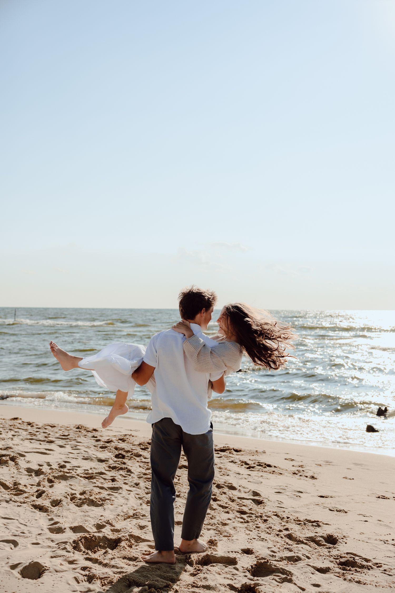 A person carries another while spinning on a beach with waves crashing in the background.