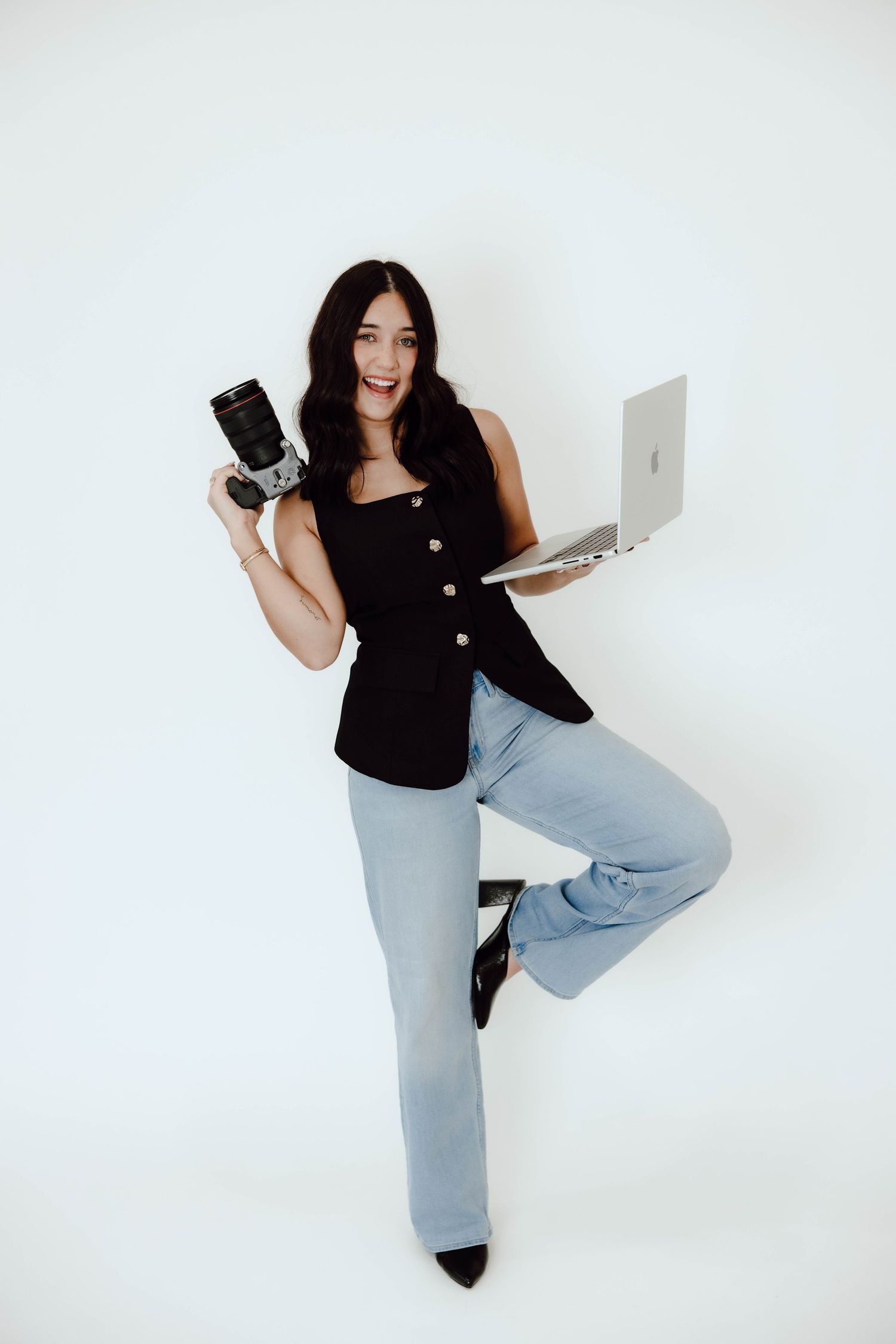 A photographer poses with camera and laptop against white background wearing black and denim.