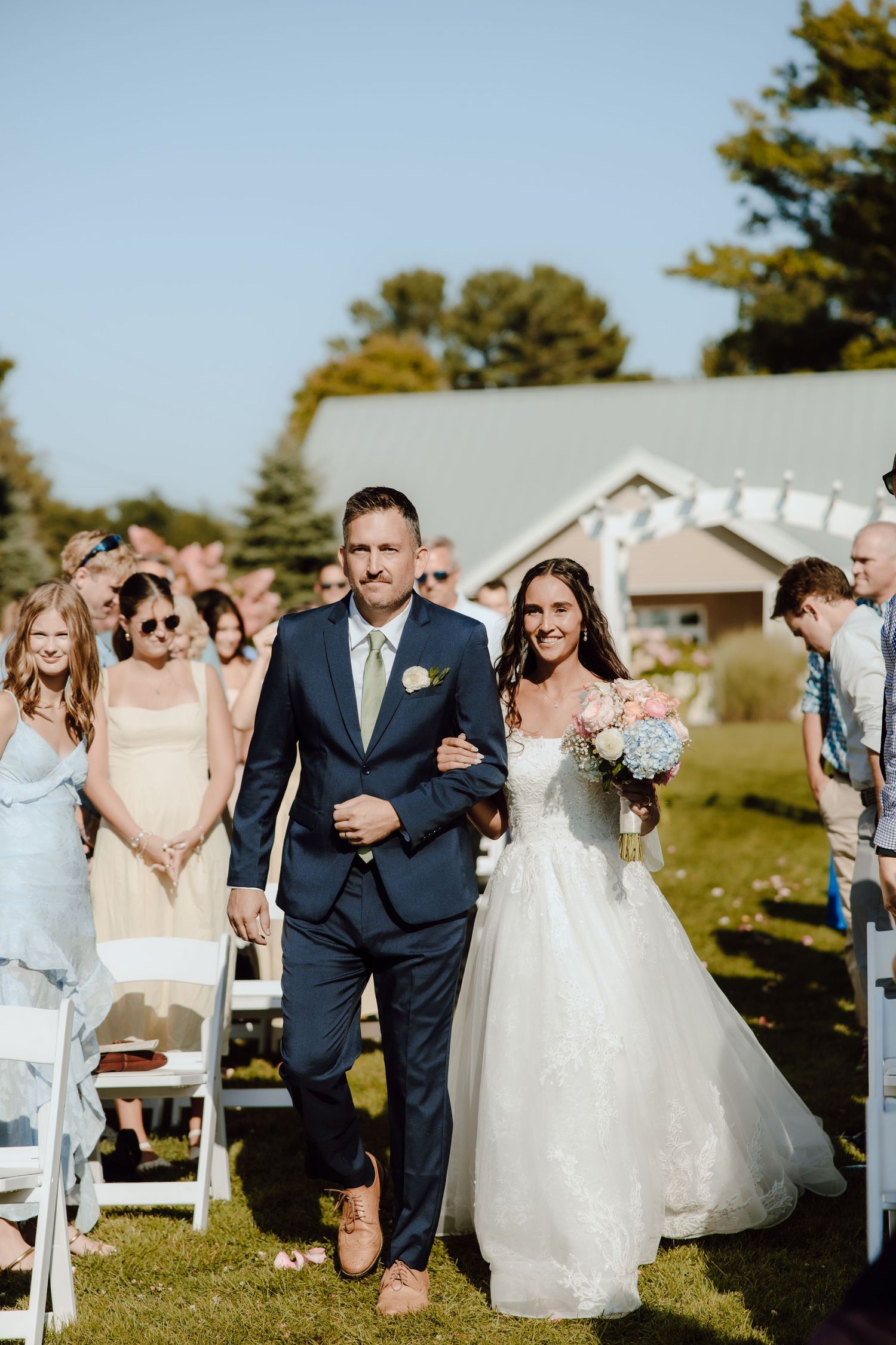 Bride in white gown walks down outdoor aisle with escort in navy suit, guests watching on sunny wedding day.
