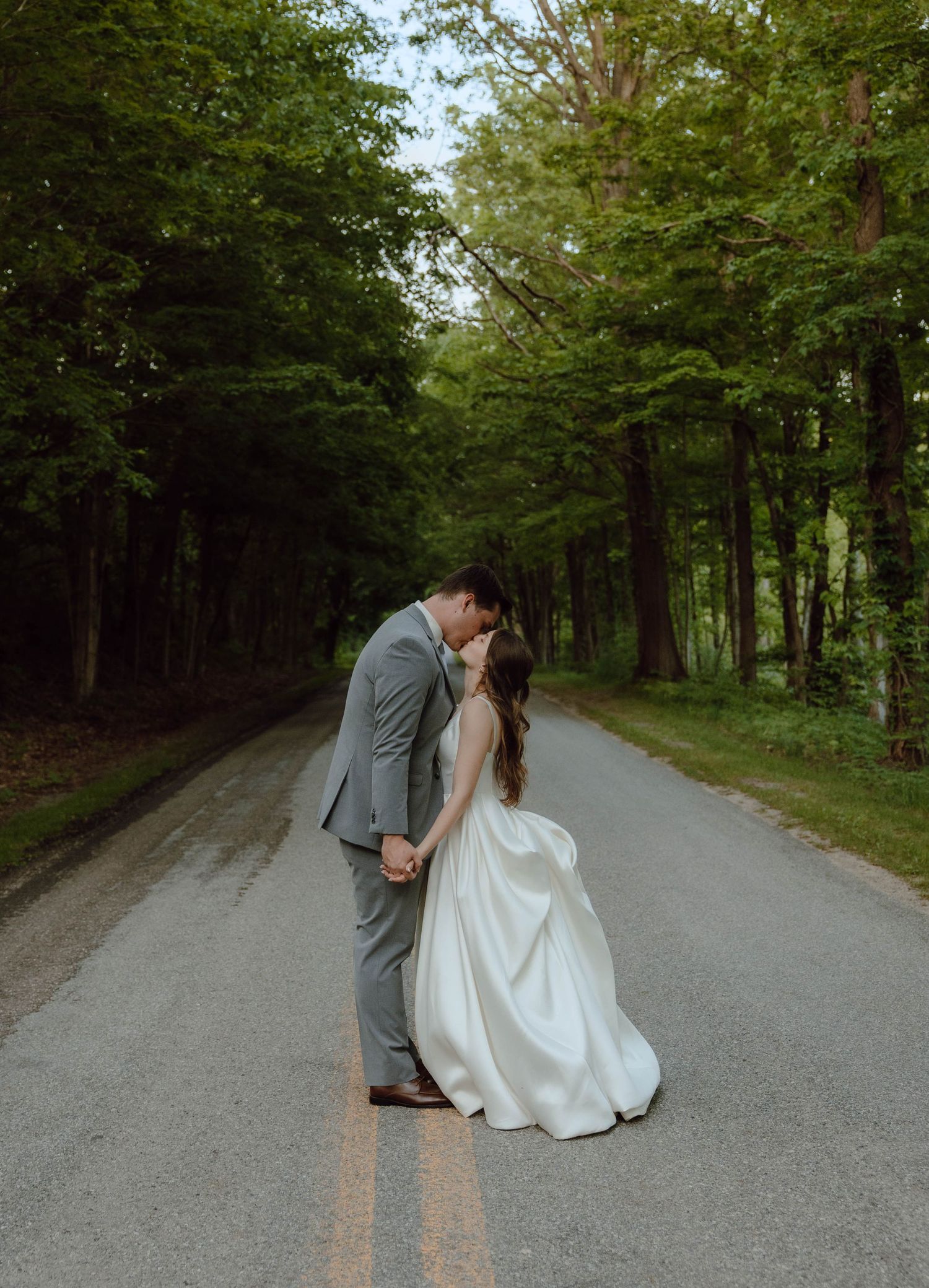 A bride and groom share a romantic kiss on a tree-lined road, bride in white gown, groom in gray suit.