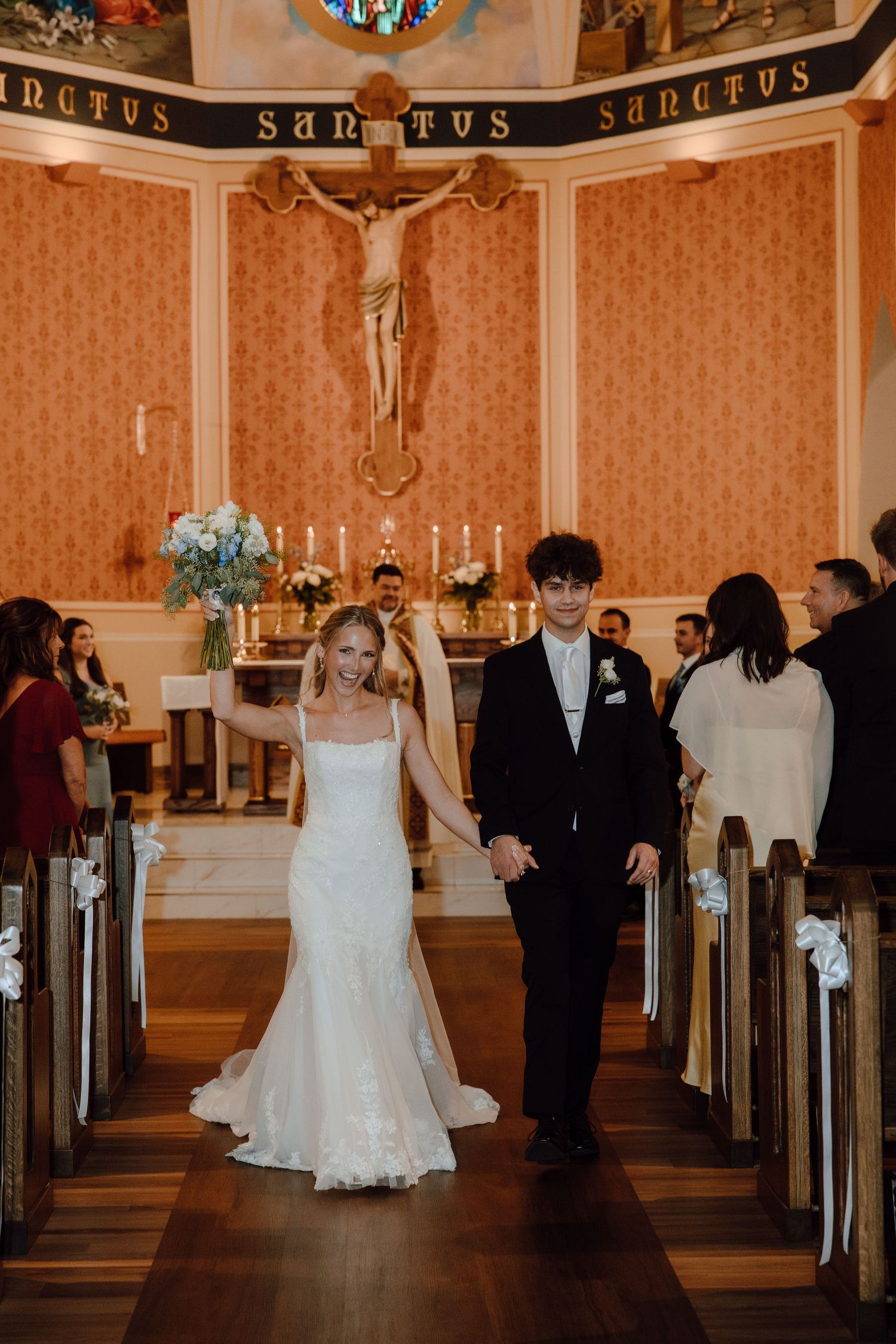 Joyful newlyweds walking back down aisle after wedding ceremony in ornate Catholic church.