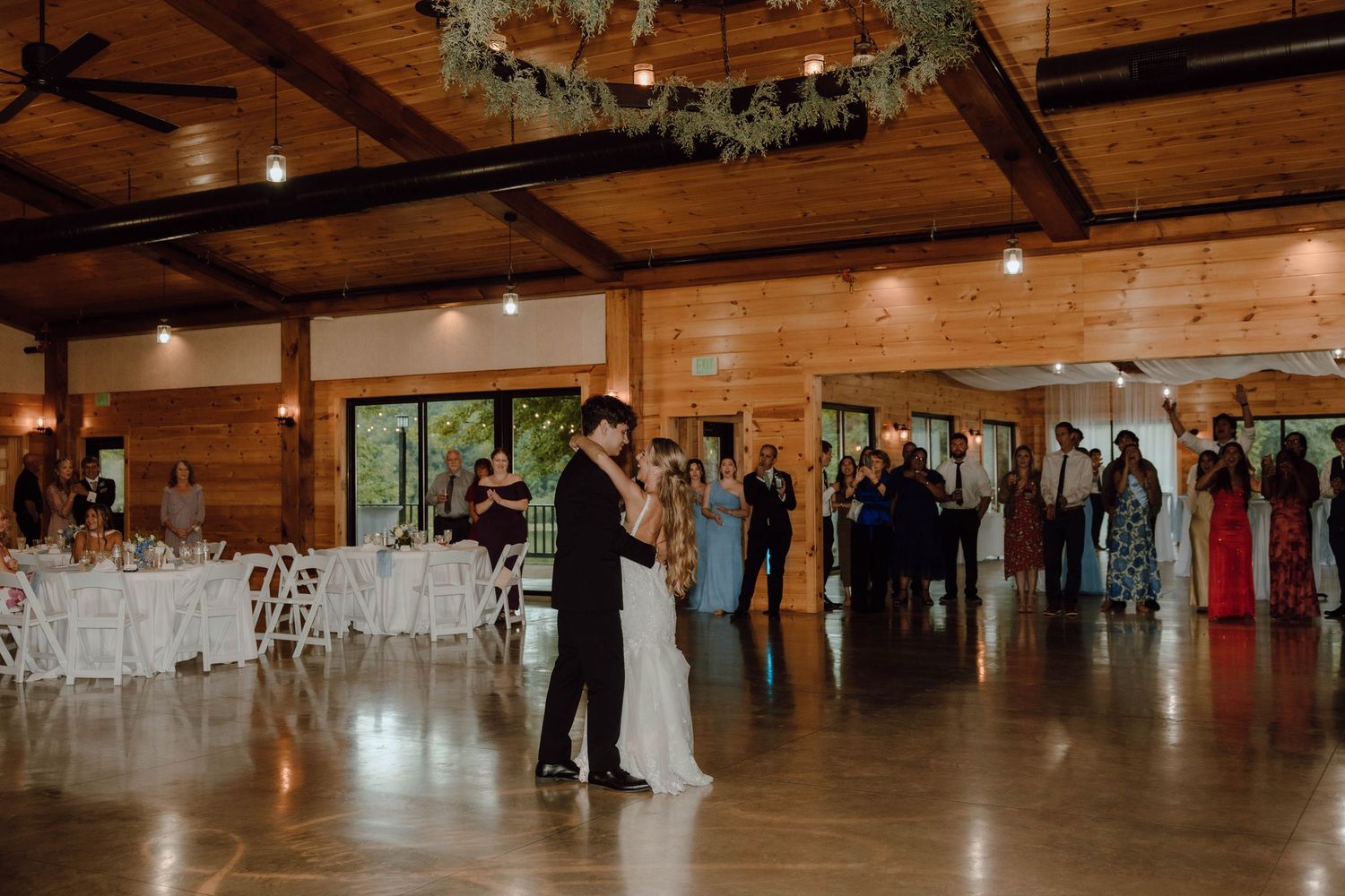 Couple shares first dance in rustic wooden wedding venue with floral chandelier and guests watching.