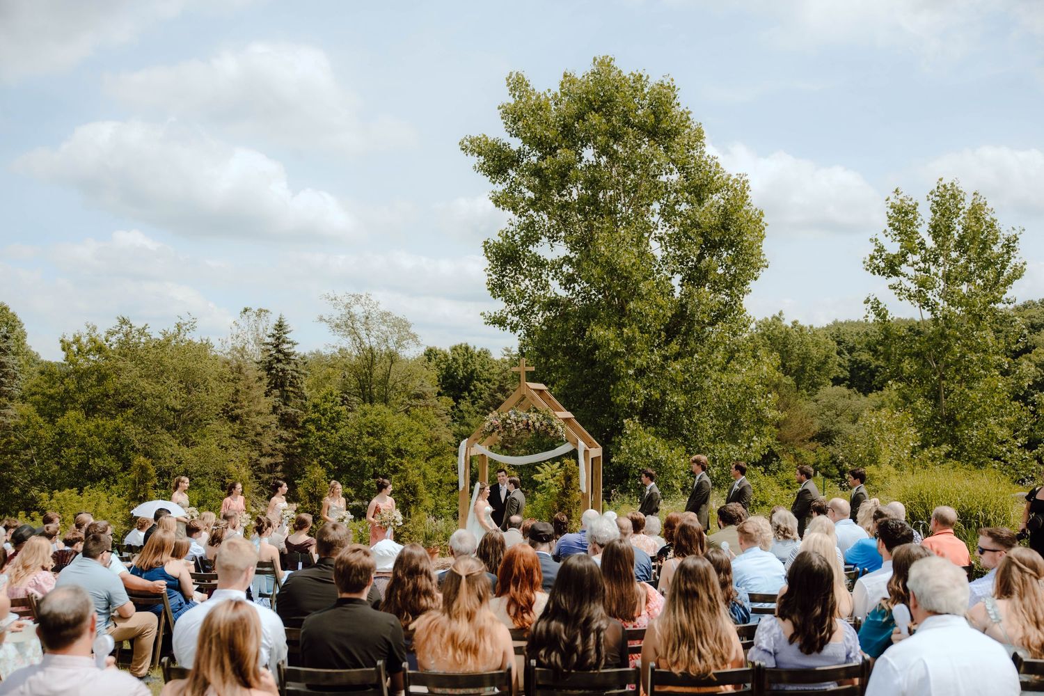 Outdoor wedding ceremony with guests seated facing couple at floral arch arbor surrounded by lush green trees under blue sky.