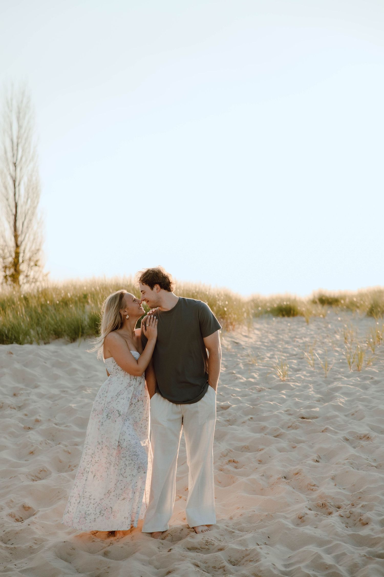 A couple shares a romantic kiss on a sandy beach path surrounded by golden dunes and green grass at sunset.