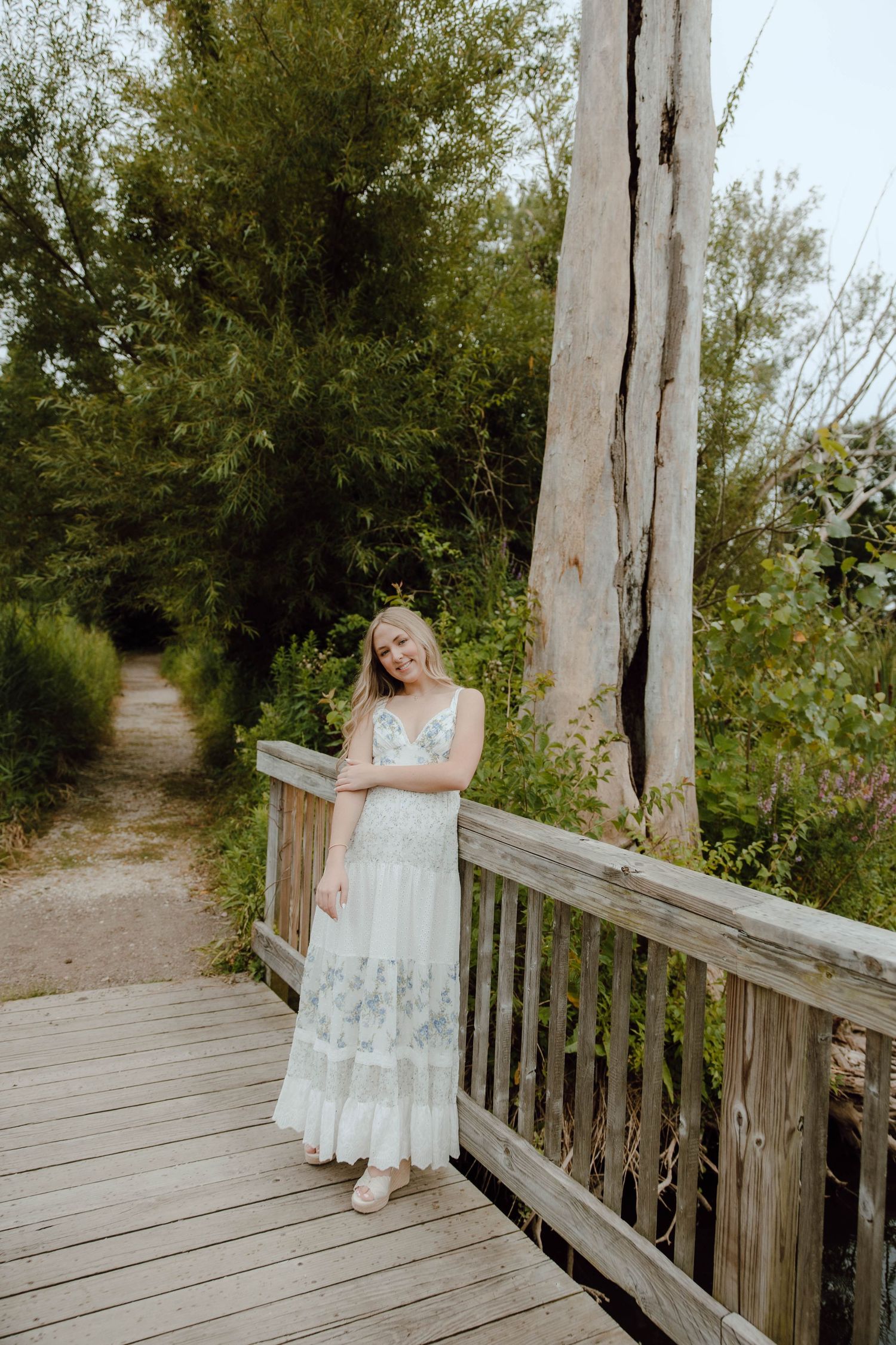 Smiling blonde woman in white floral maxi dress posing on wooden bridge surrounded by green trees.