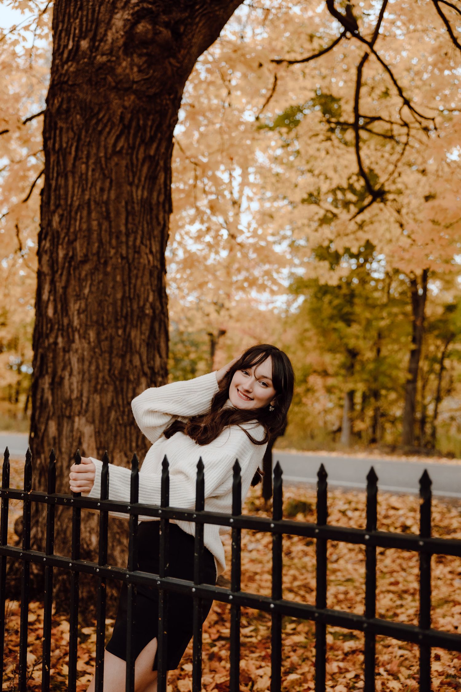 Person in white sweater poses playfully on black iron fence surrounded by fall foliage.