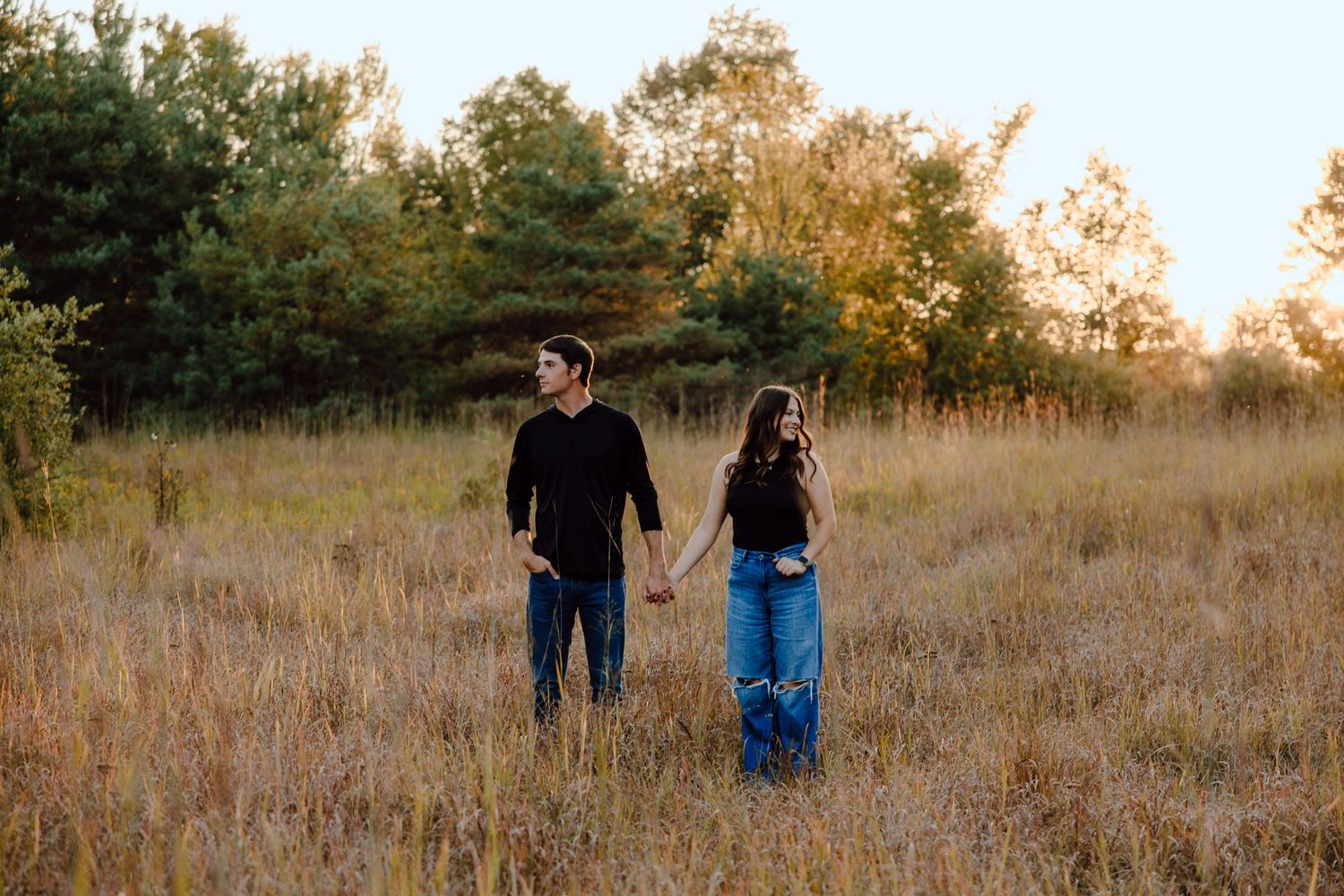 Couple walking together through a golden field at sunset with trees in the background.