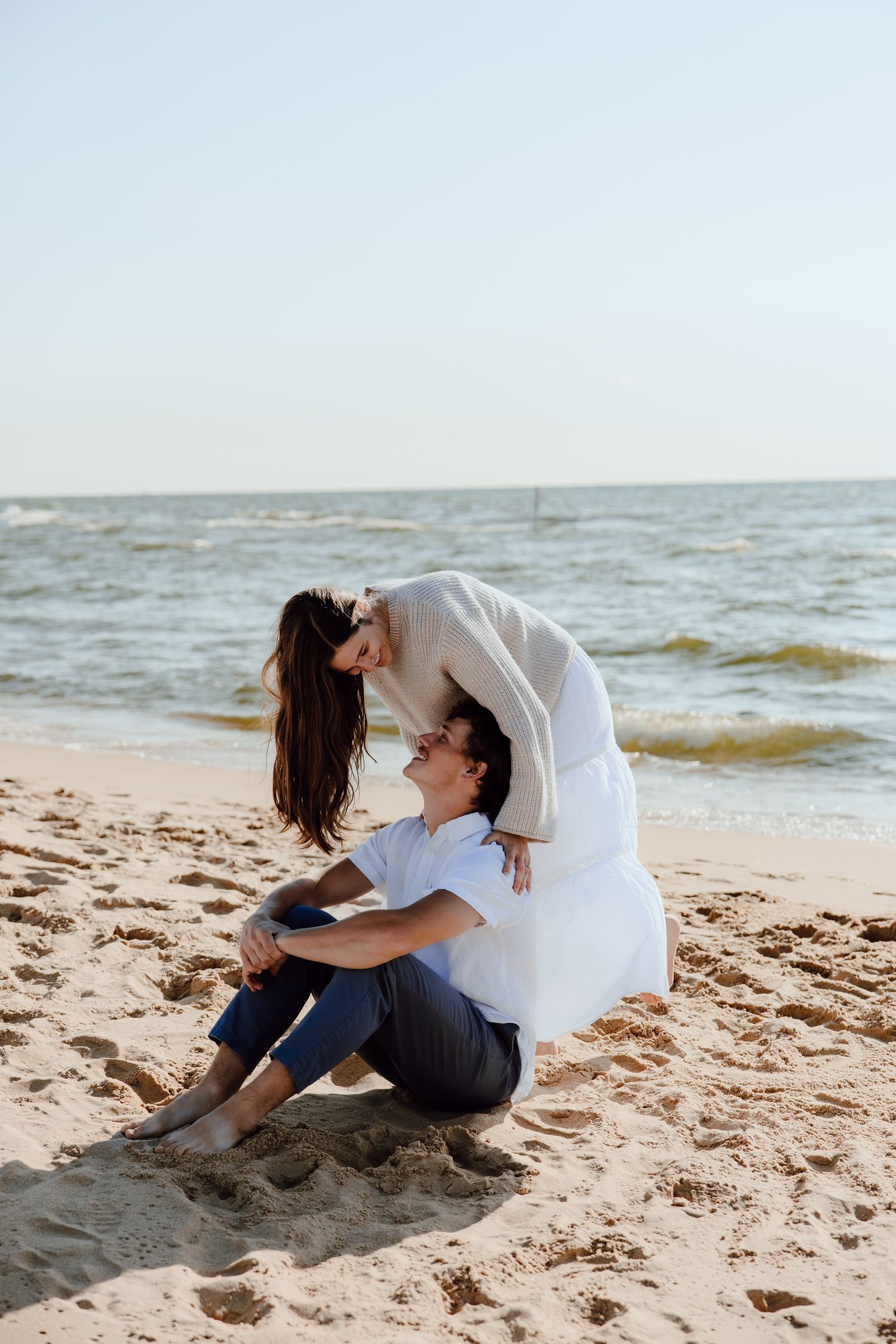 A playful couple poses on the beach as one person leans over the other sitting in the sand.