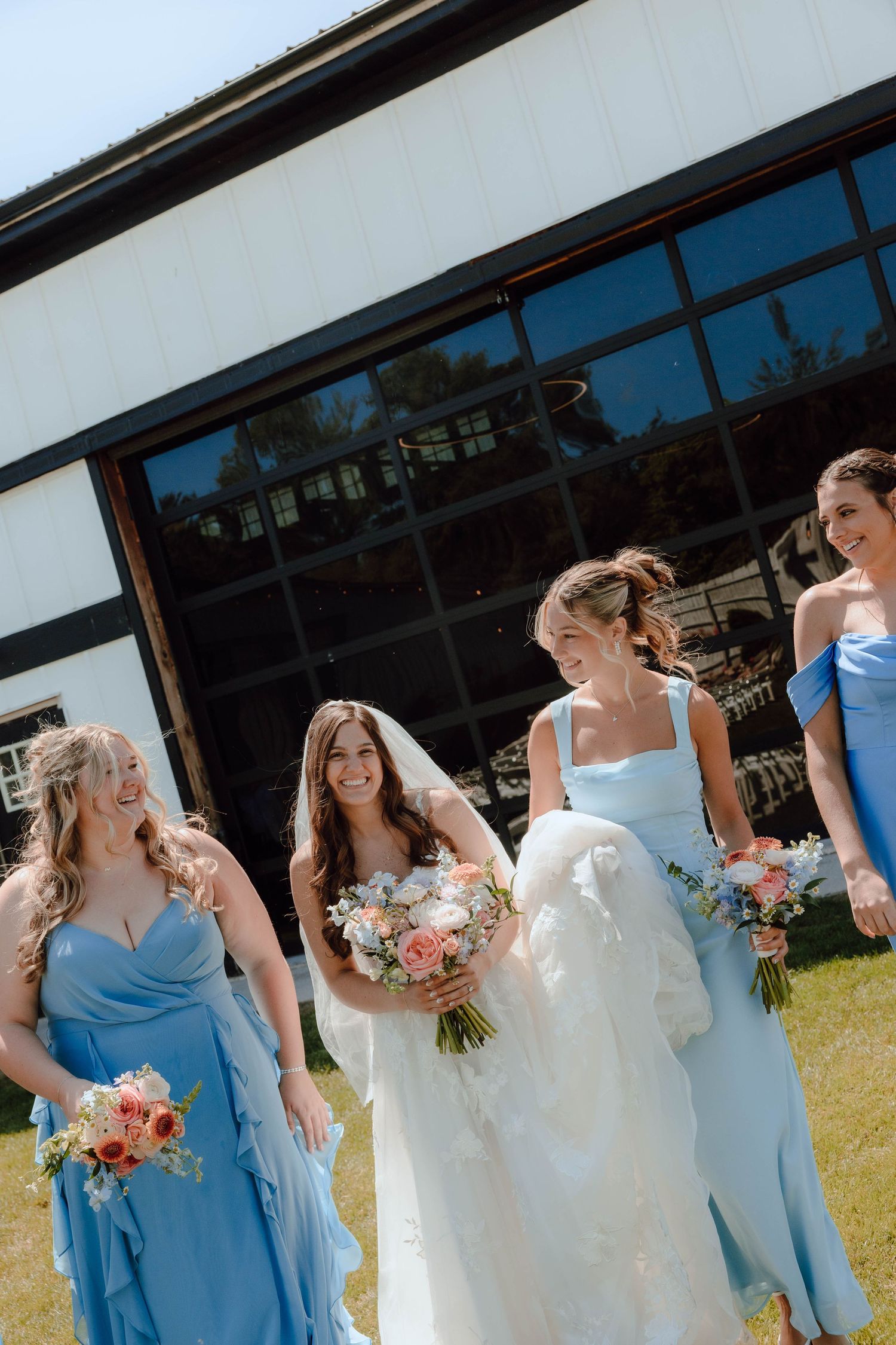 Bride in white gown with bridesmaids in blue dresses holding bouquets, smiling outdoors near a barn.