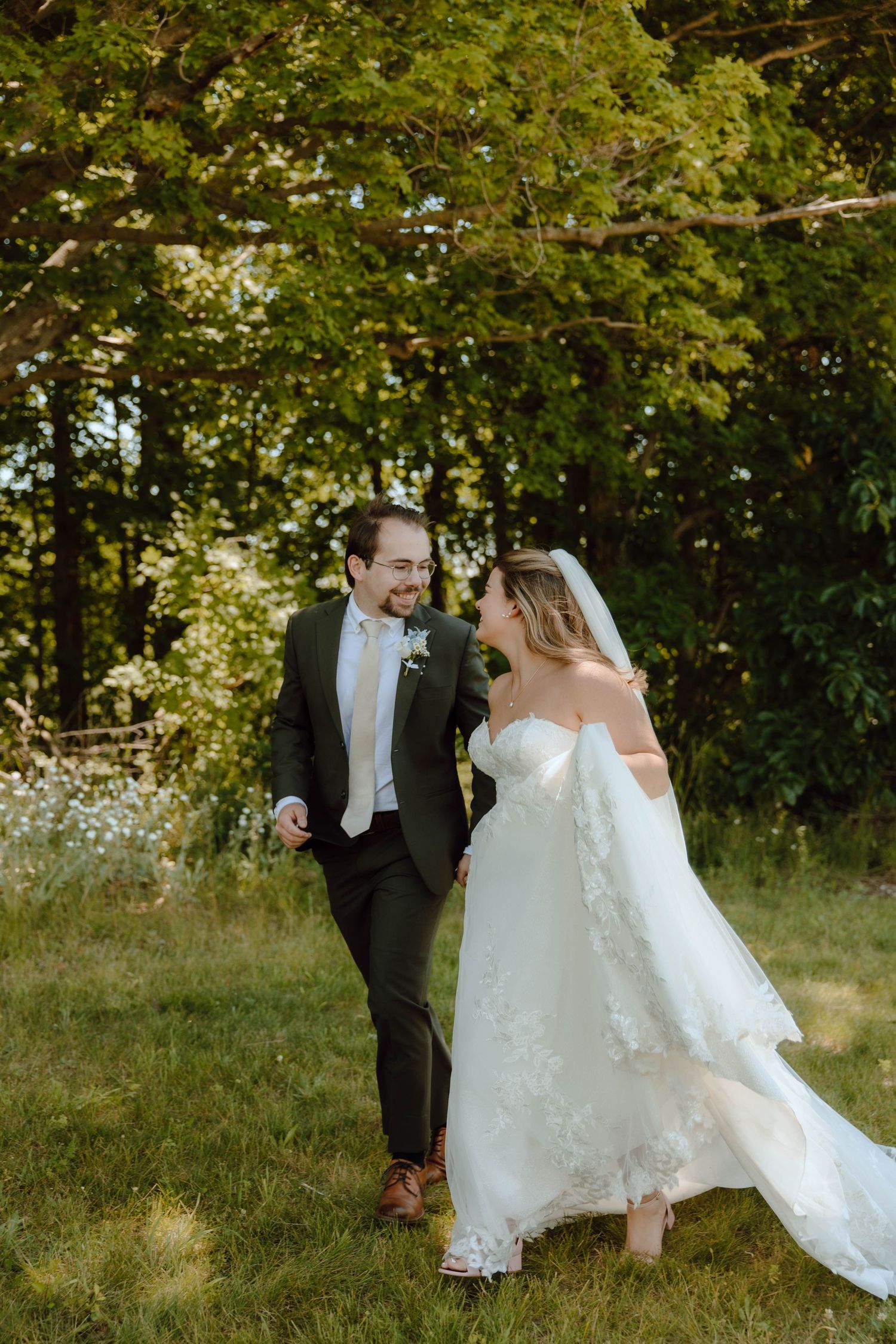 A bride and groom walk hand in hand through a sunlit garden, smiling at each other in elegant wedding attire.