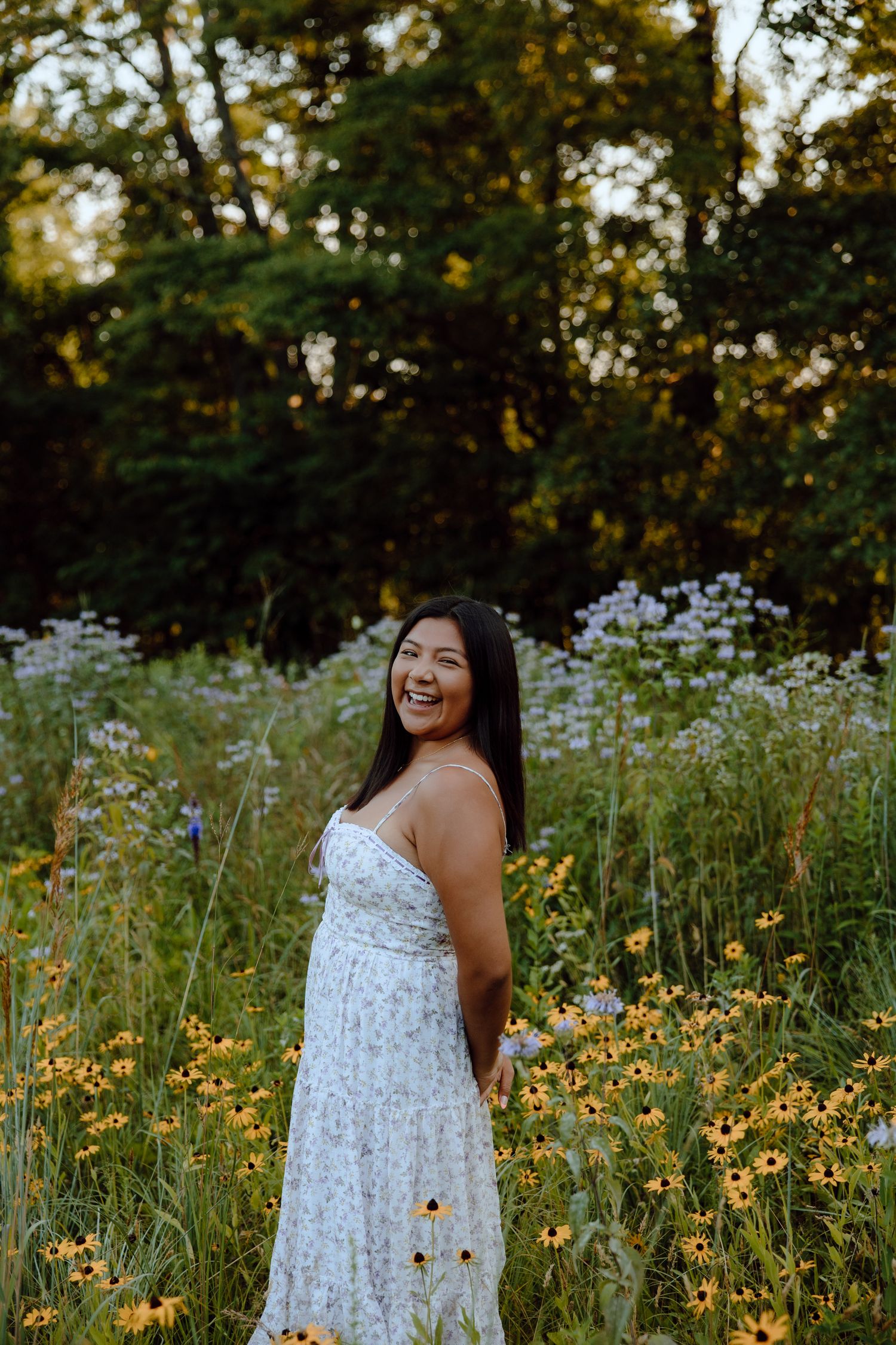 A smiling woman in a floral white dress stands among yellow wildflowers and greenery in a sunlit outdoor field.