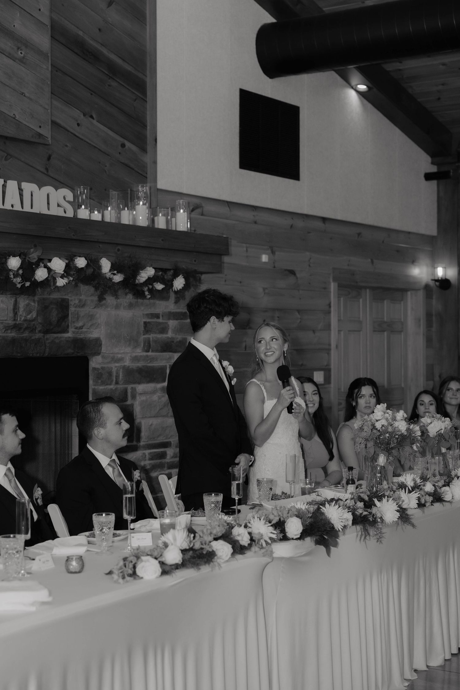 Newlyweds giving speech at elegant rustic wedding reception with floral centerpieces.