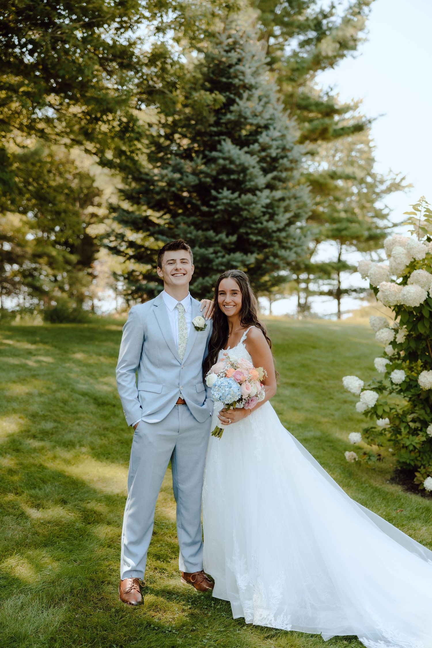 Smiling bride and groom pose together outdoors, bride holding a pastel bouquet, groom in a light blue suit.