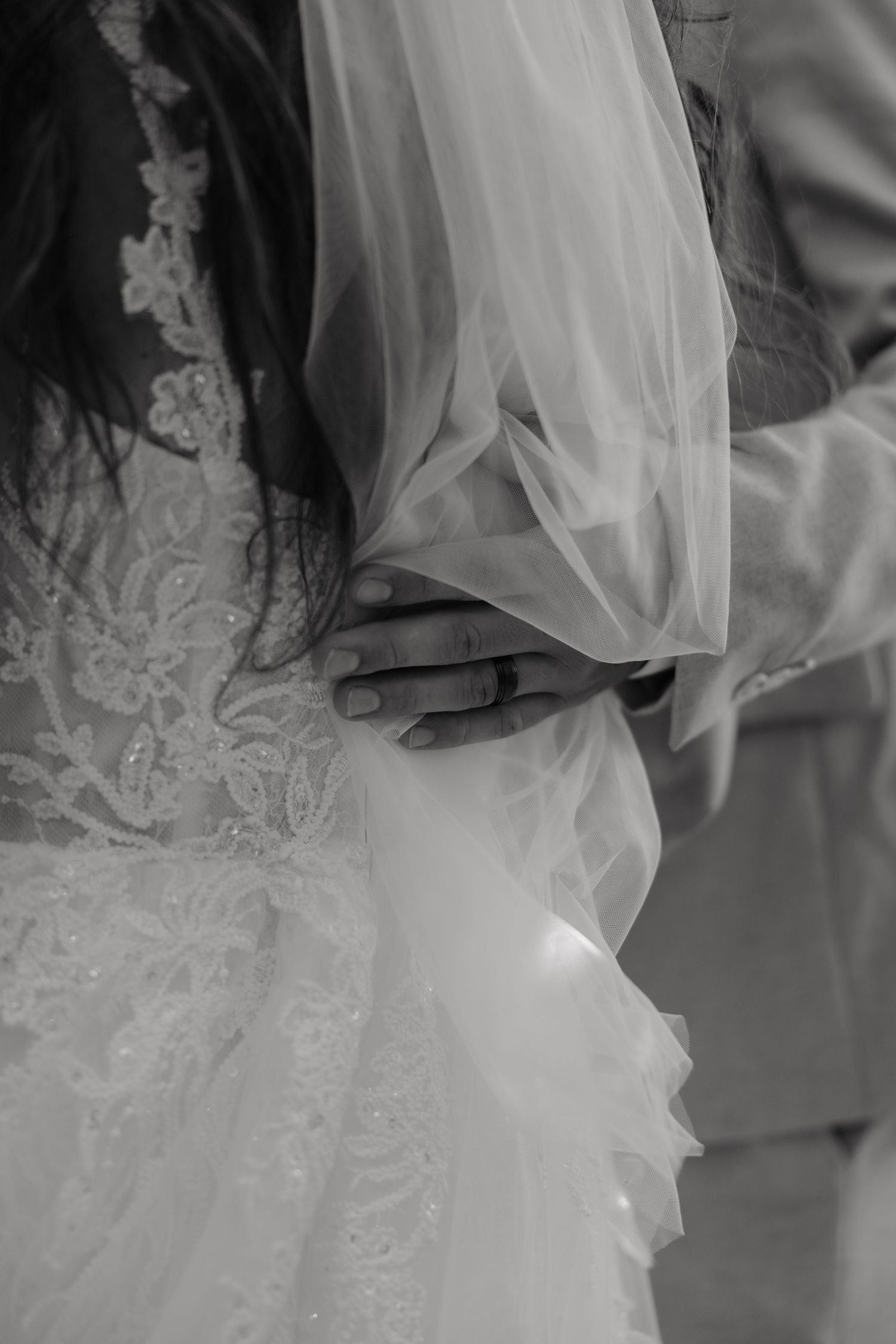 Black and white close-up of groom's hand on bride's lace wedding dress waist with veil.