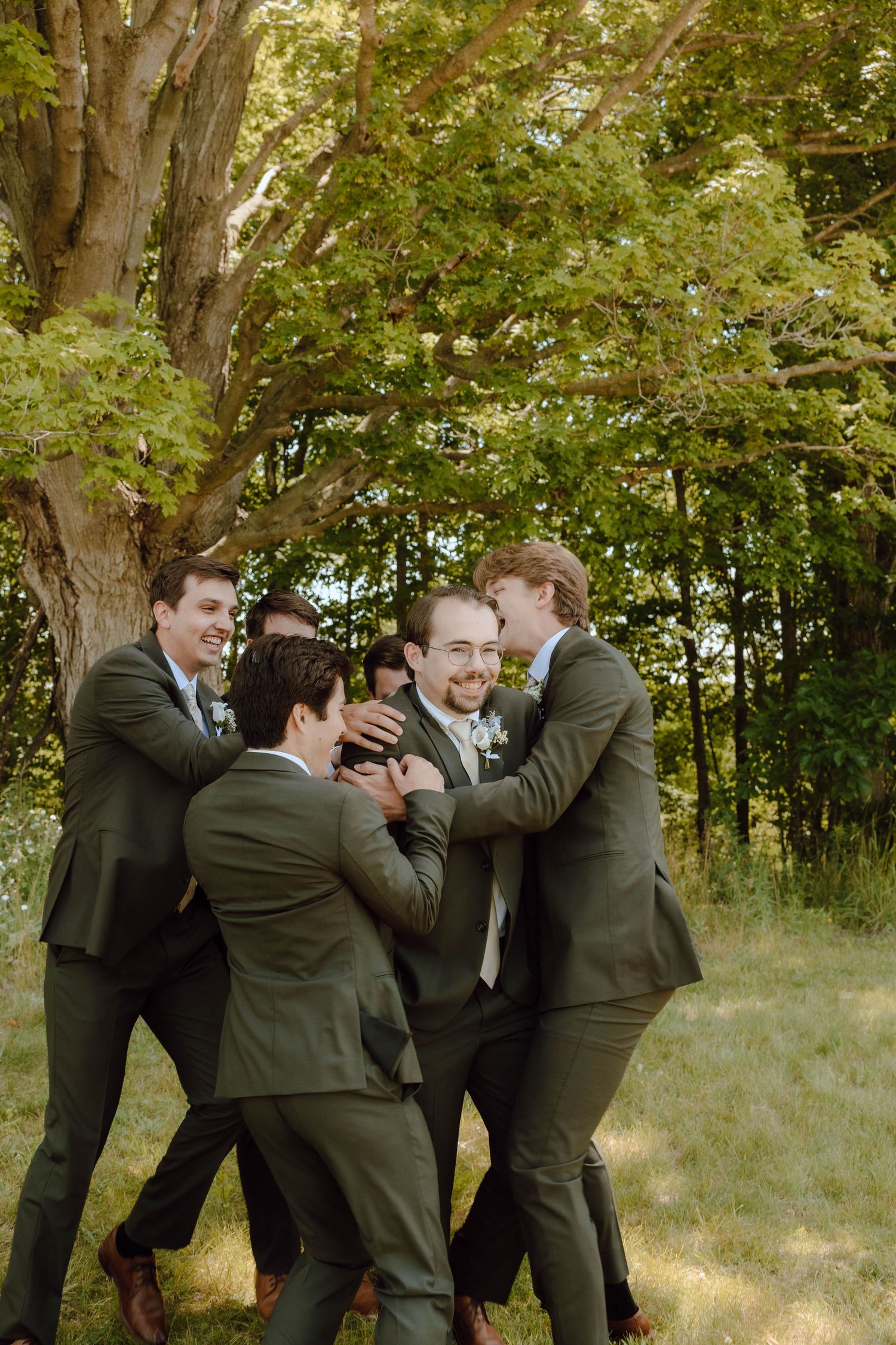 Groom laughing with groomsmen in olive suits celebrating outdoors near large tree.