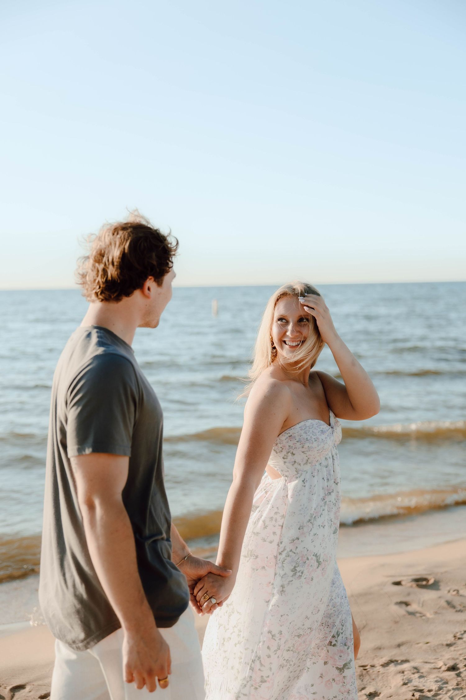 A smiling blonde woman in a floral dress holds hands with a man in a gray shirt on a sunny beach.