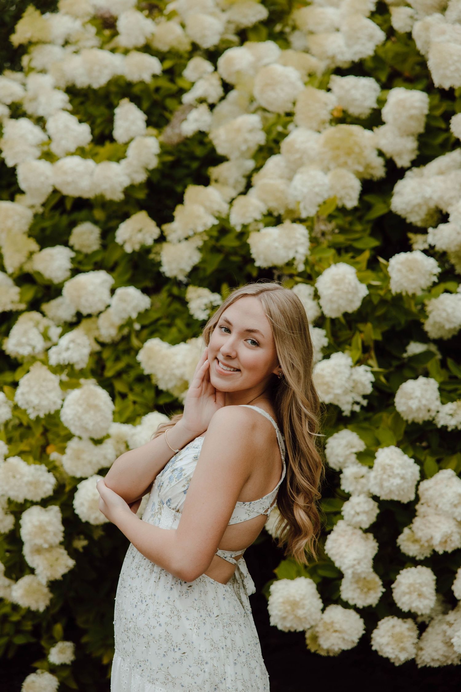 Young woman in floral dress smiling in front of lush white hydrangea blooms in a garden setting.