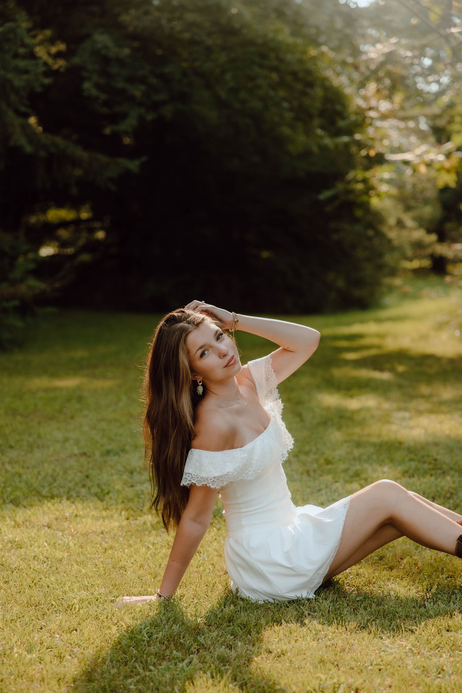 Young woman in white off-shoulder dress sitting on green grass in a sunlit garden, posing gracefully.