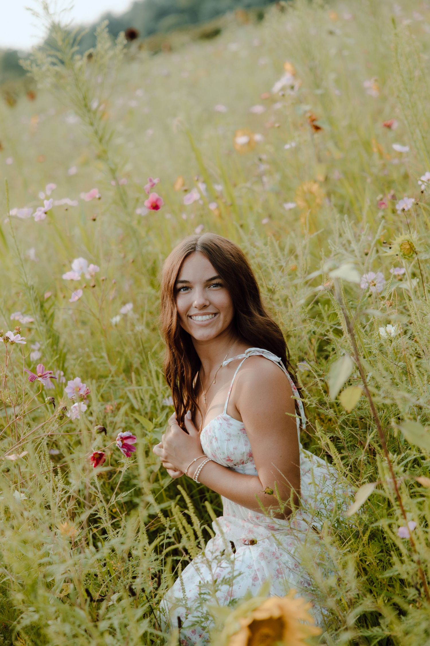 Young woman in floral dress smiling in a wildflower meadow with pink and yellow blooms on a sunny day.