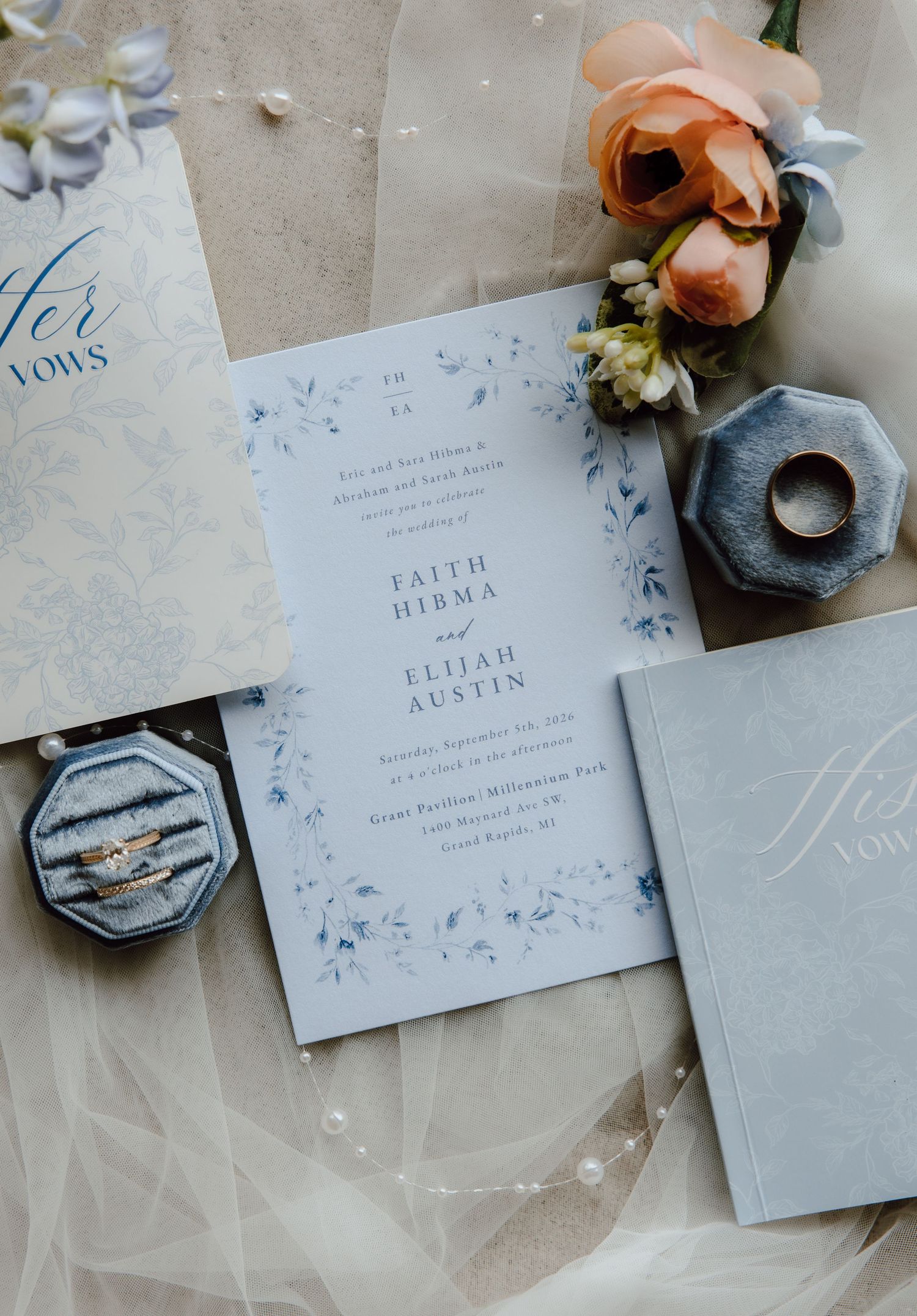 Elegant wedding invitation suite with dusty blue floral details, velvet ring box, gold bands, and peach roses on soft tulle.