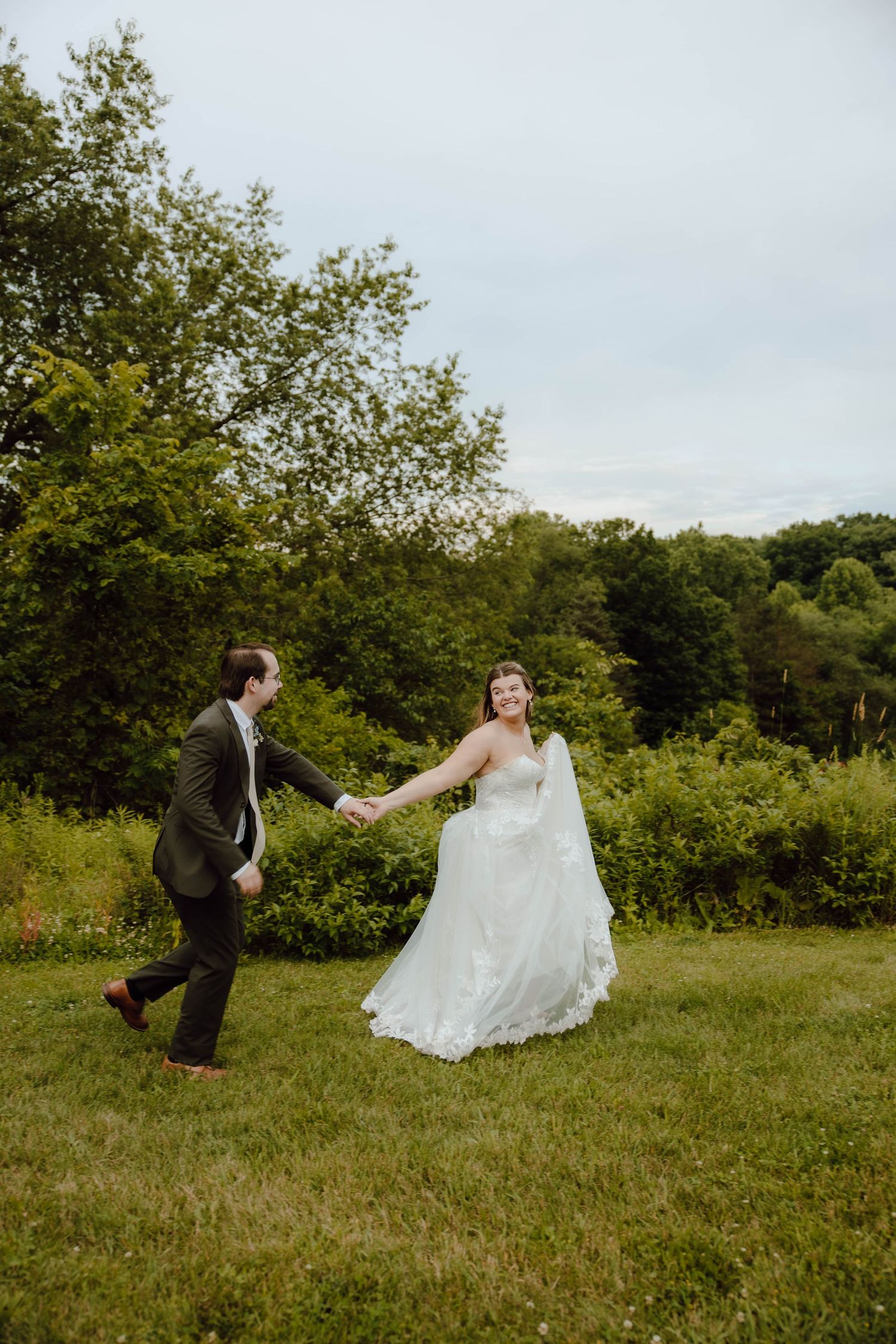 A joyful bride and groom hold hands while walking through a lush green meadow on their wedding day.