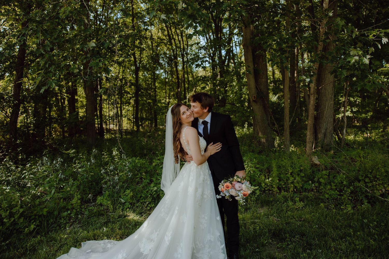 Bride and groom share a romantic kiss in a lush green forest, bride wearing a flowing white gown with veil.