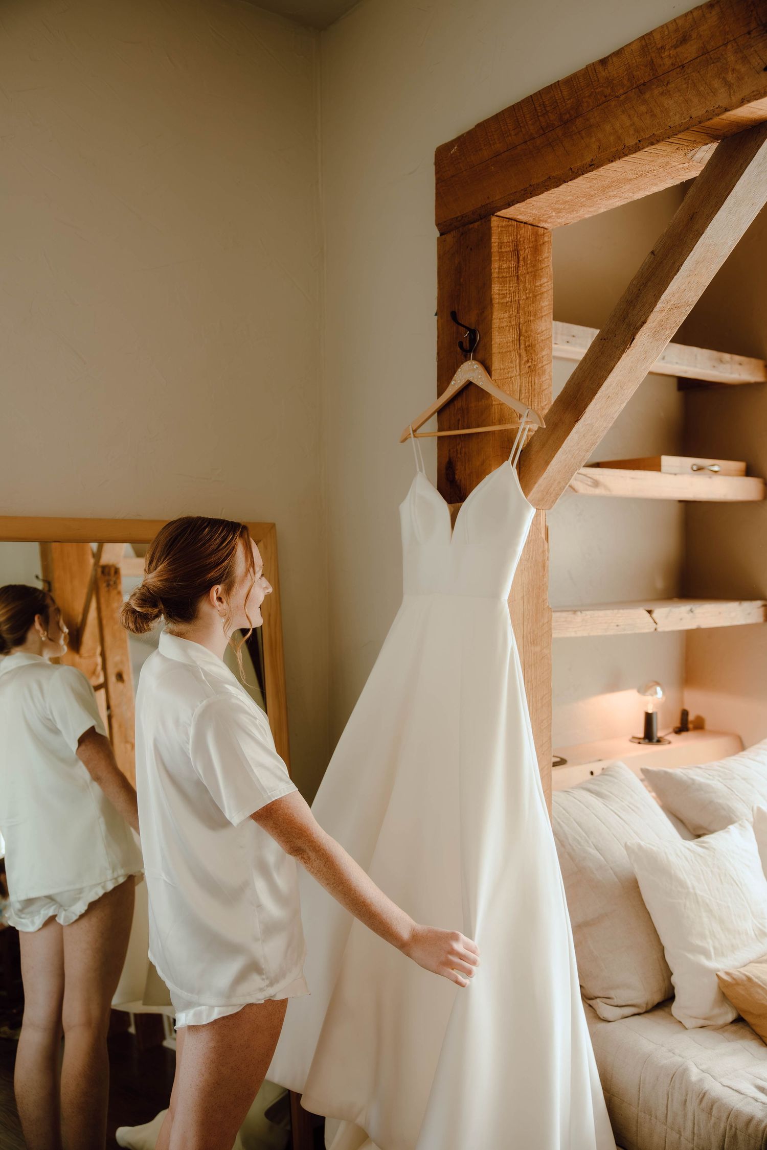 Bride in white robe admiring her wedding dress hanging on a rustic wooden beam in a cozy room.