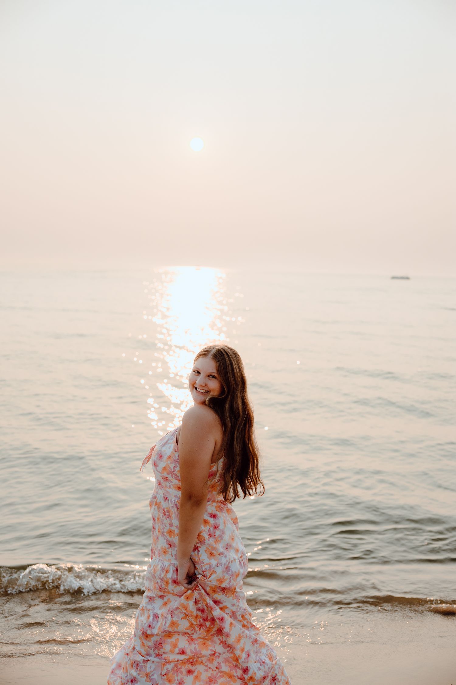 Person in floral dress twirls on beach at sunset with ocean waves in background.