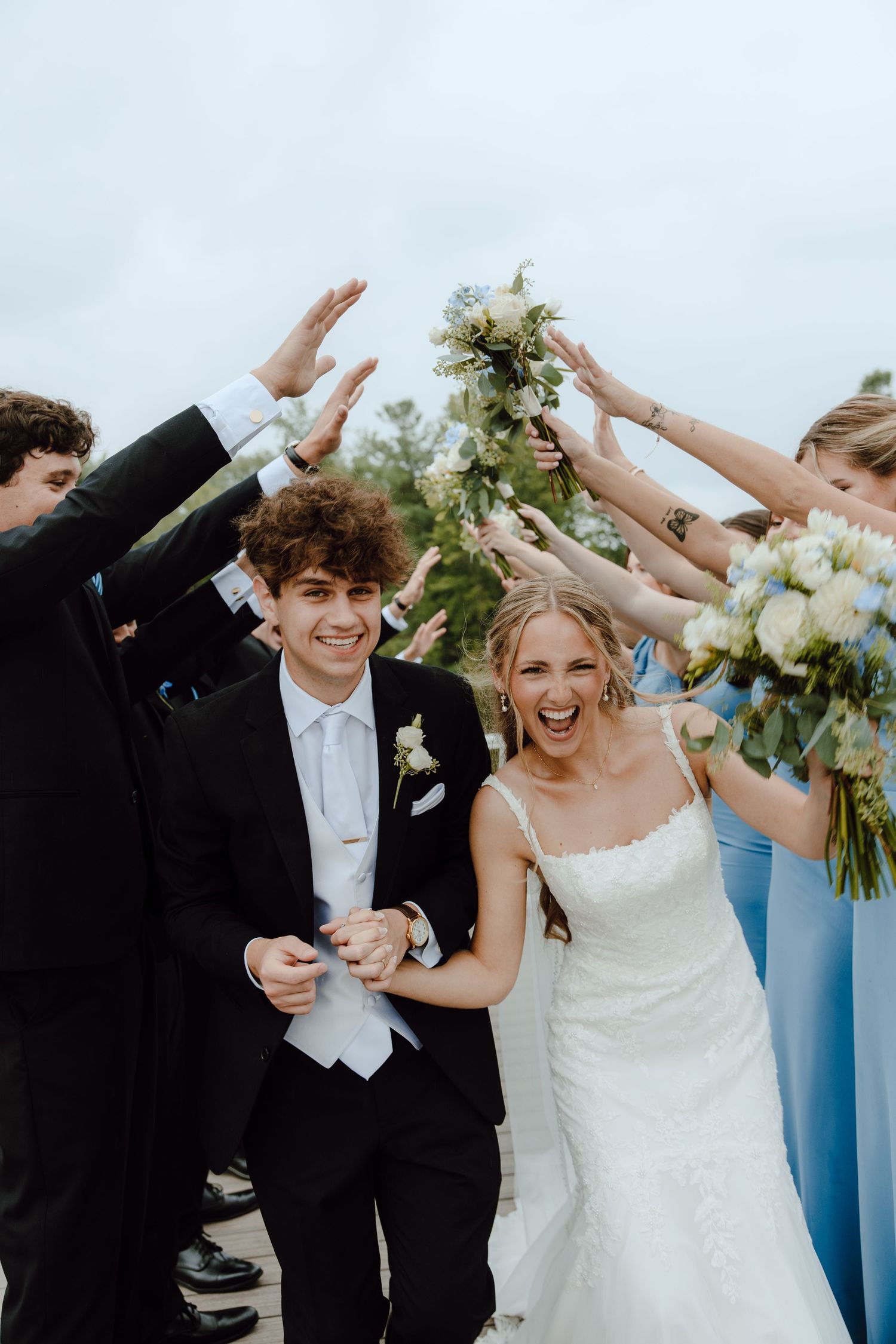 Joyful newlyweds walk through an arch of raised hands formed by wedding guests after their outdoor ceremony.