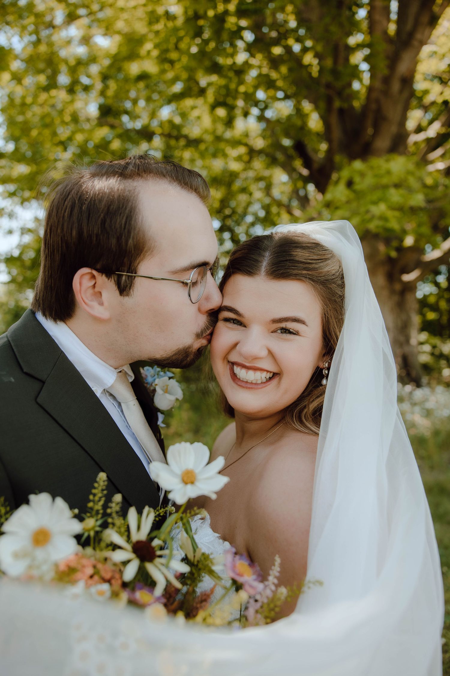 A groom kisses his smiling bride on the cheek as she holds a wildflower bouquet outdoors on their wedding day.
