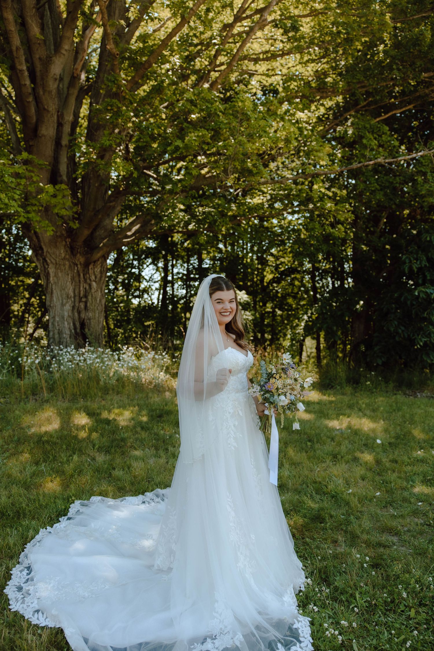 Smiling bride in lace wedding gown and veil holding bouquet outdoors among green trees.