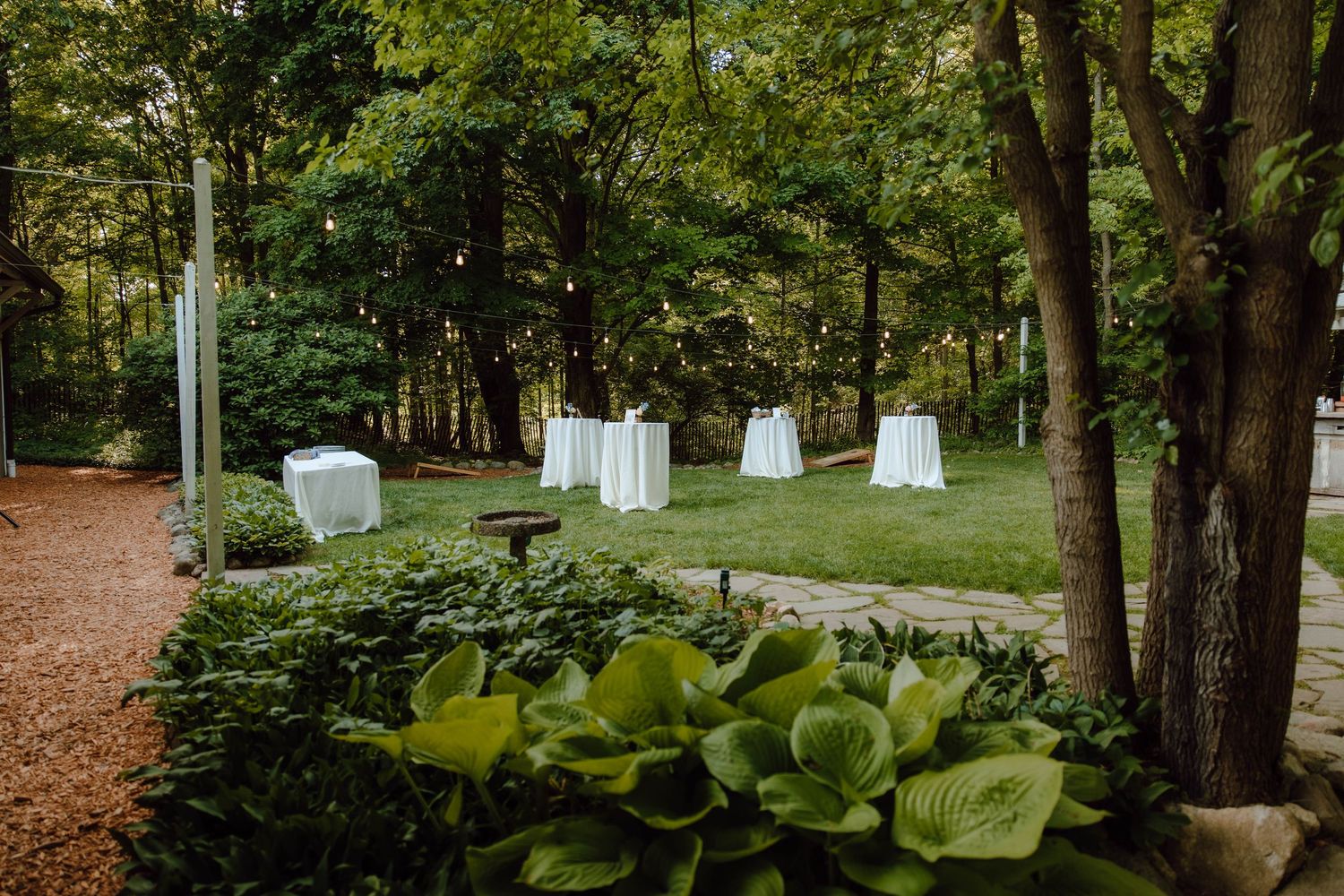 Elegant outdoor cocktail hour setup with white draped high-top tables arranged on a garden lawn.
