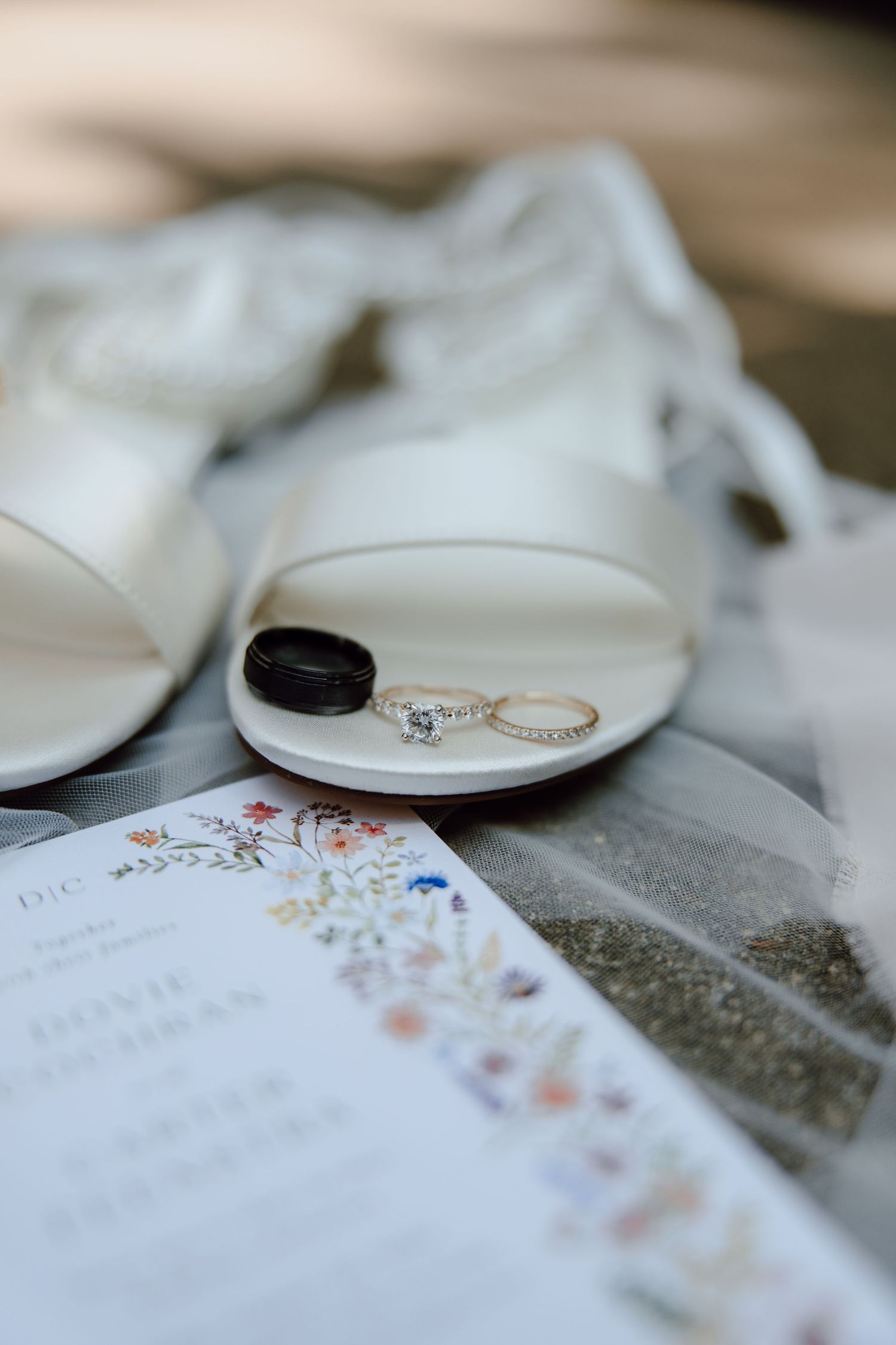 Wedding rings and bands displayed on a white dish beside bridal heels and floral invitation.