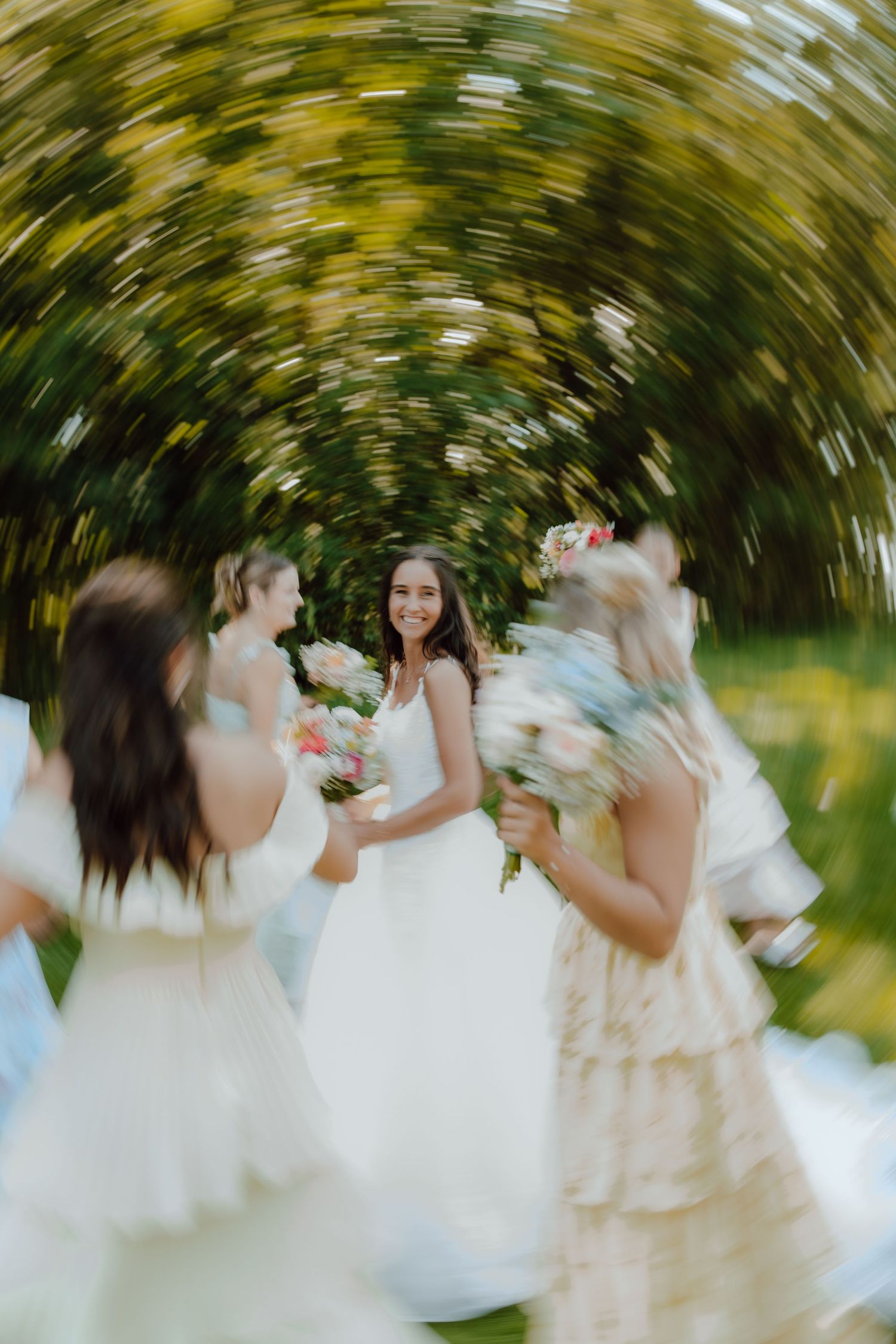 Bride smiling with bridesmaids holding bouquets outdoors with artistic spin blur effect.