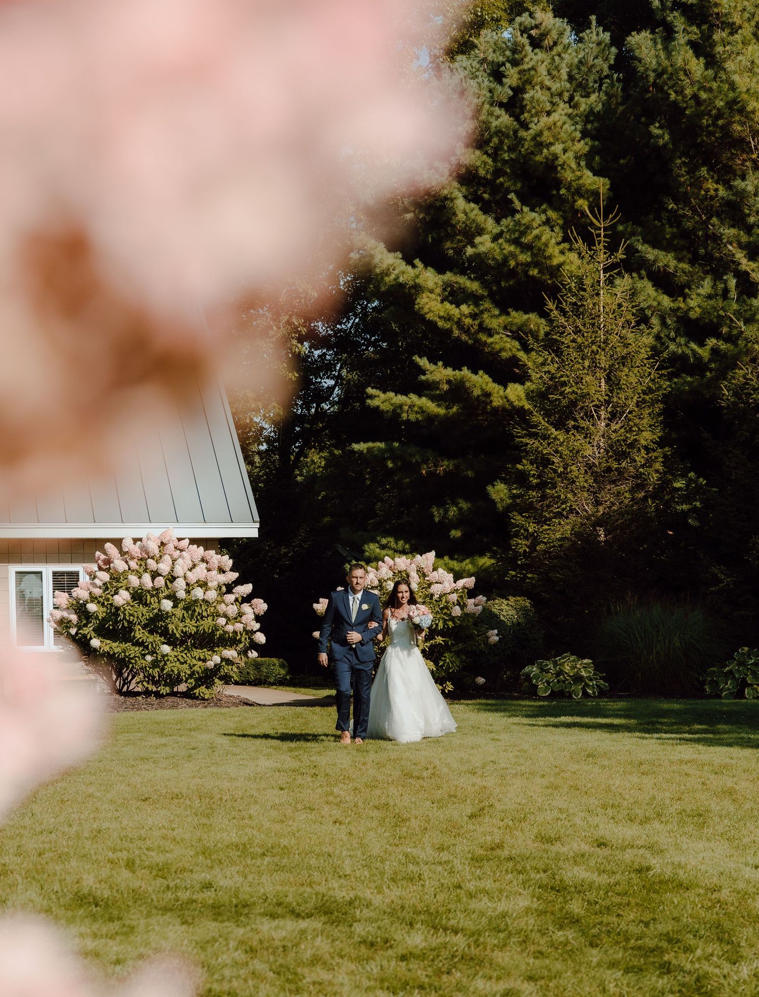 Bride and groom walking on lawn framed by pink blooms and hydrangeas near white building.